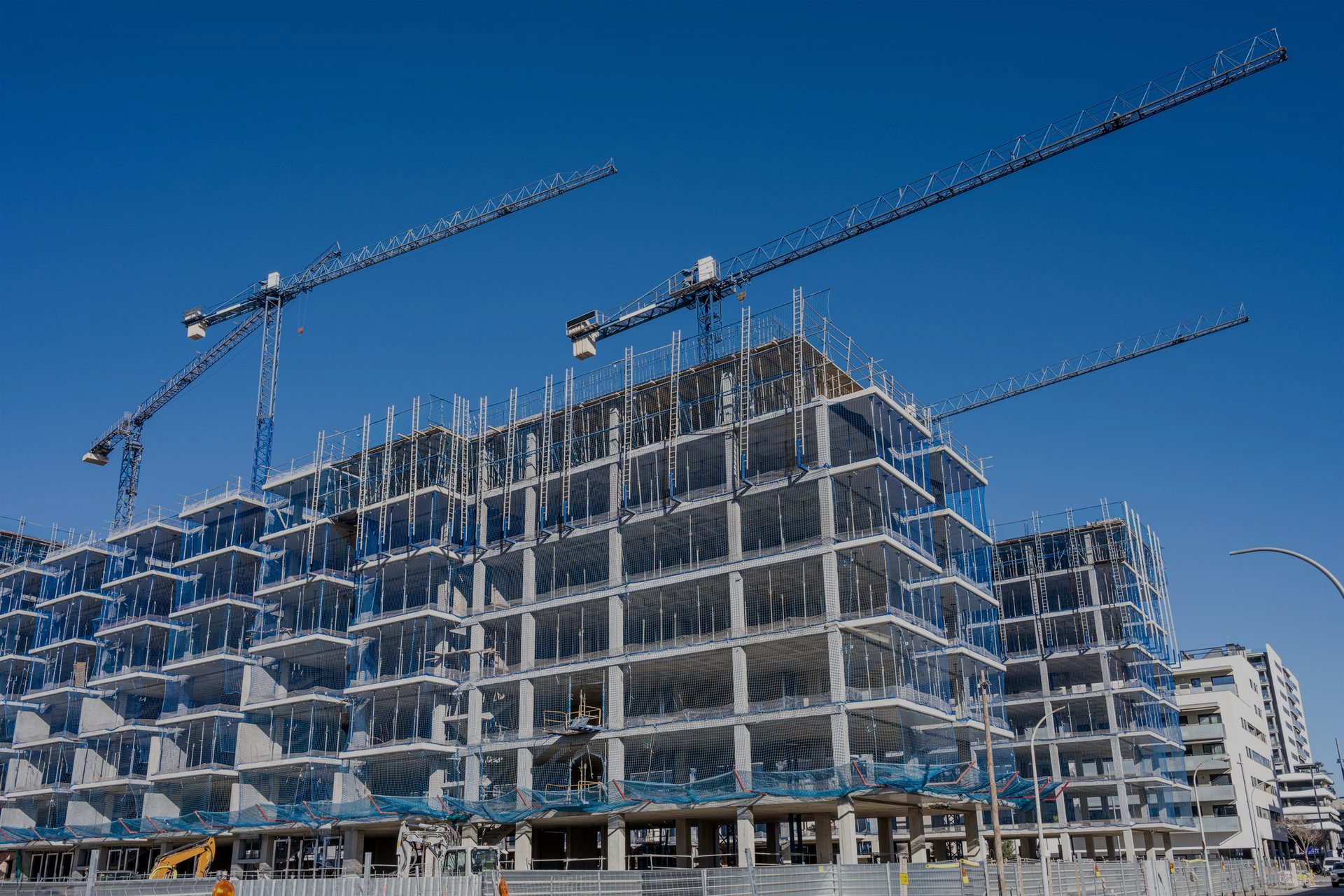 Cantiere di un edificio a più piani sotto un cielo azzurro e terso, con tre gru in alto.