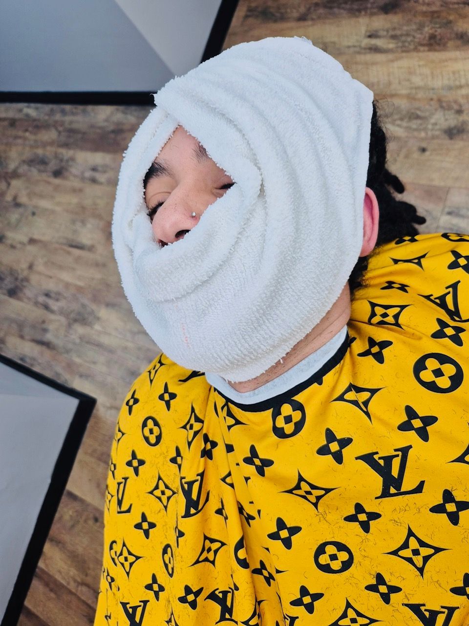 Person with a white towel wrapped around face, wearing a yellow Louis Vuitton print shirt.