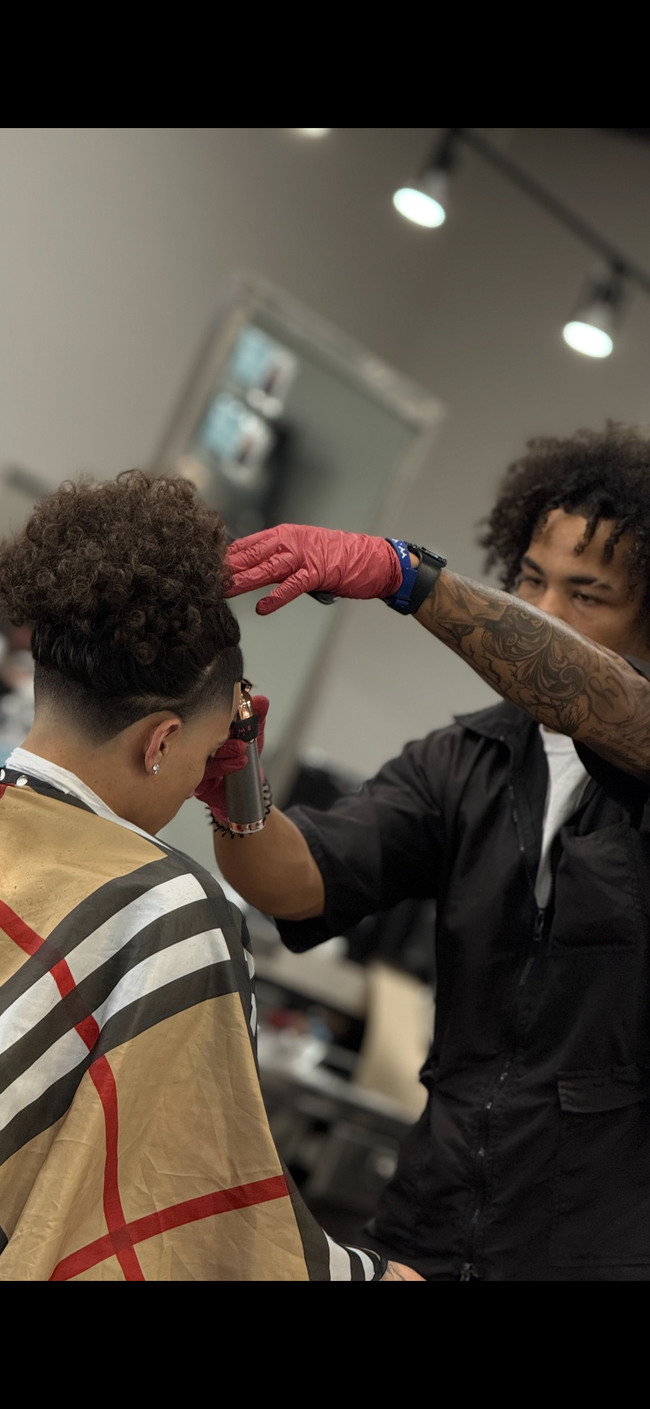 A barber cutting a client's hair in a salon. The client wears a Burberry patterned cape. The barber has tattoos.
