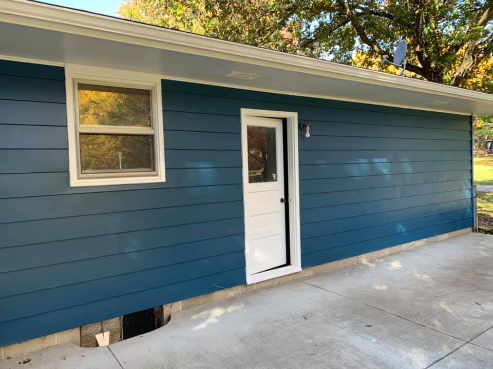 Blue-sided building with a white door and window, set on a concrete slab; blue siding, white trim.