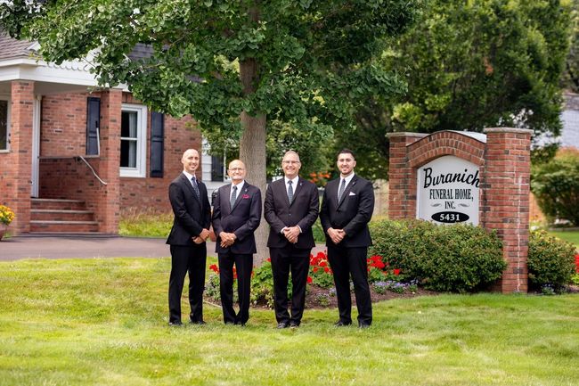 Four men in suits stand outside a brick building with a sign that reads