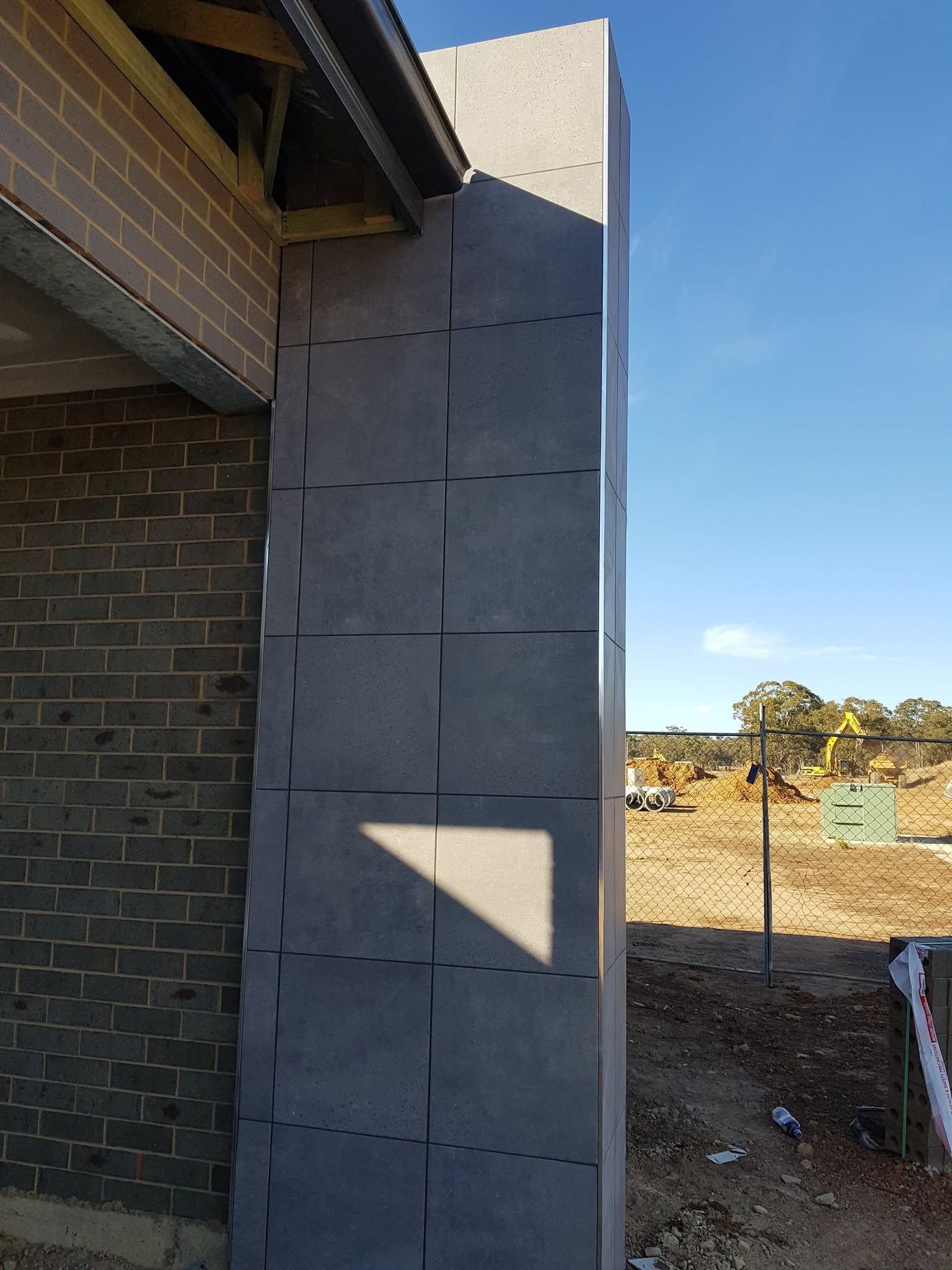 Exterior wall under construction with gray sheathing beside a brick house and dirt lot under blue sky