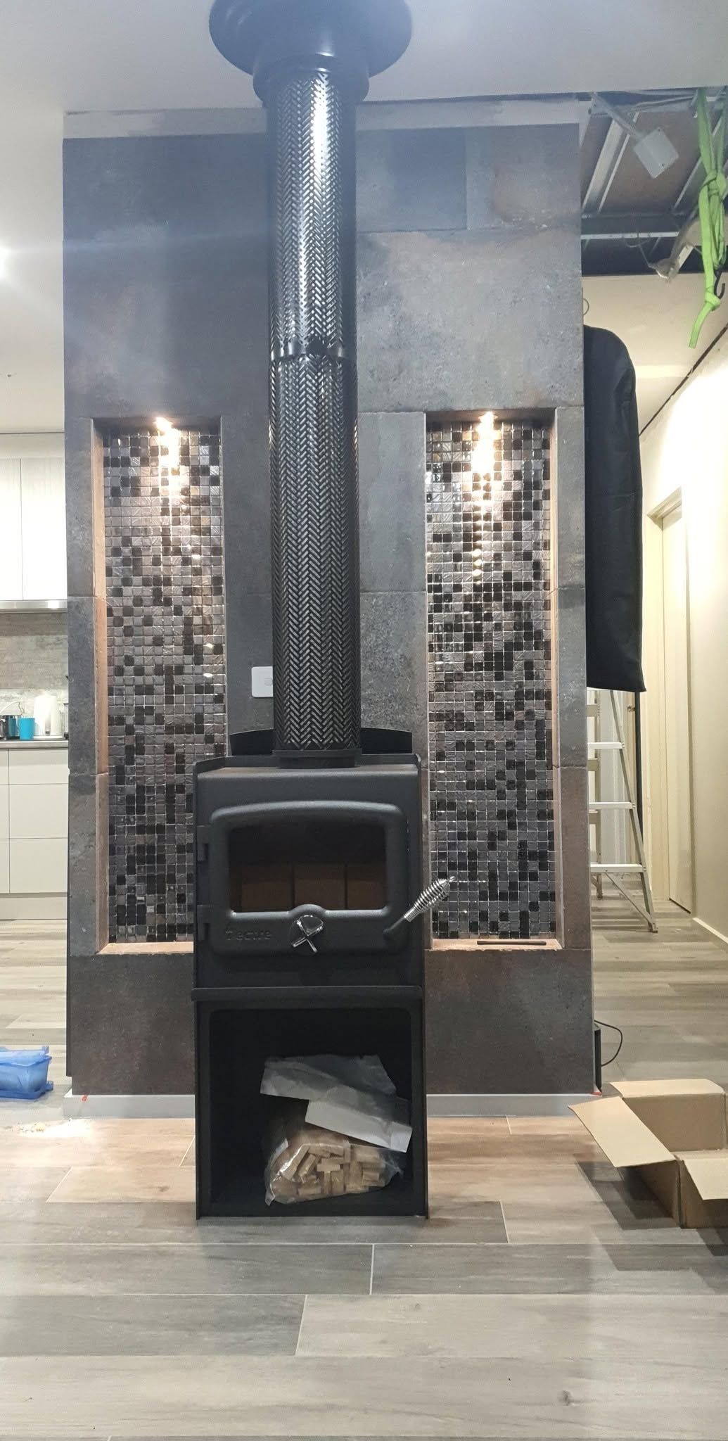 Decorative black wood stove with tall chimney against a brick-pattern wall in a tiled indoor space