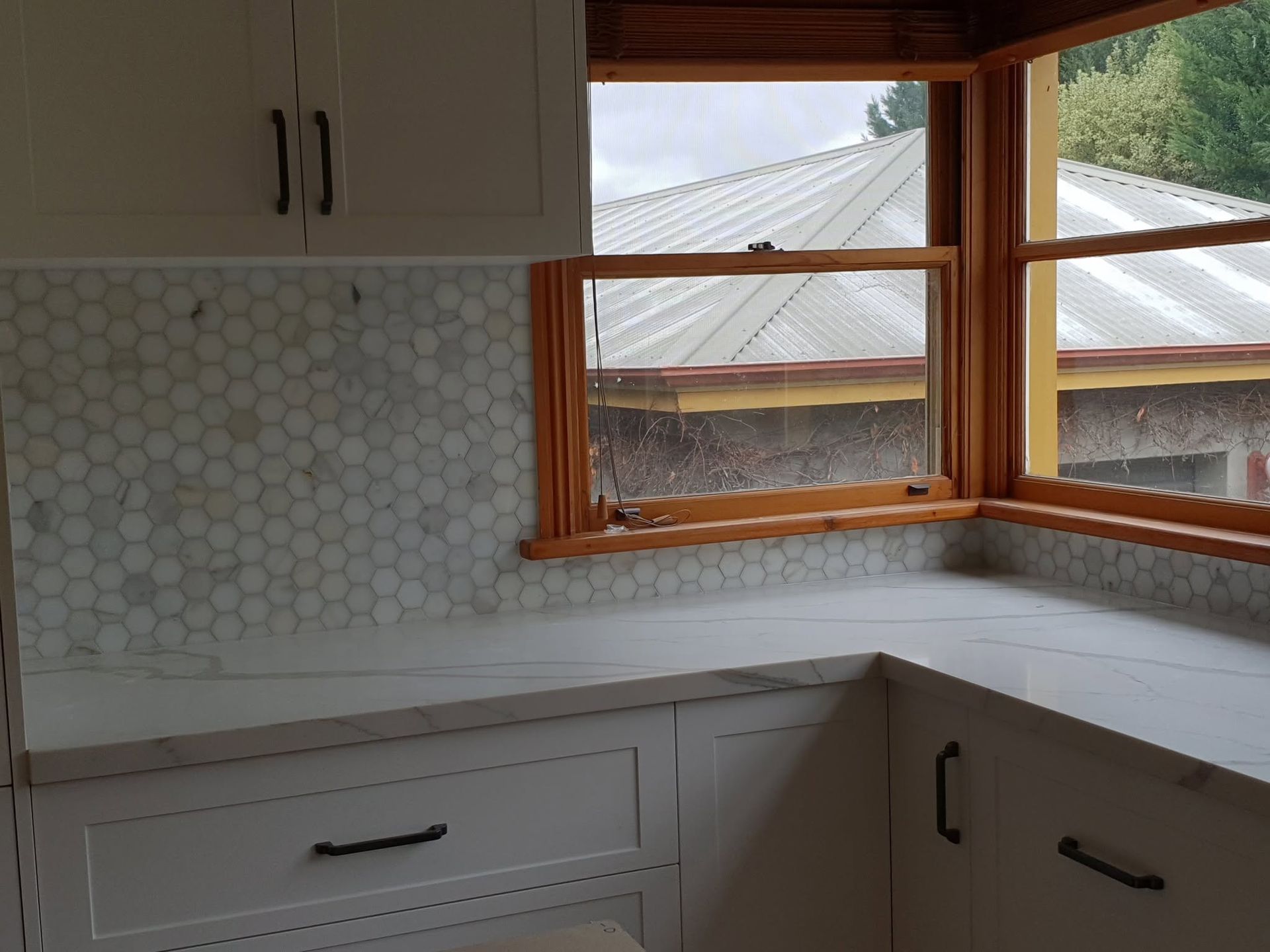 Empty kitchen corner with white cabinets, wood-trimmed windows, and a light hex tile backsplash