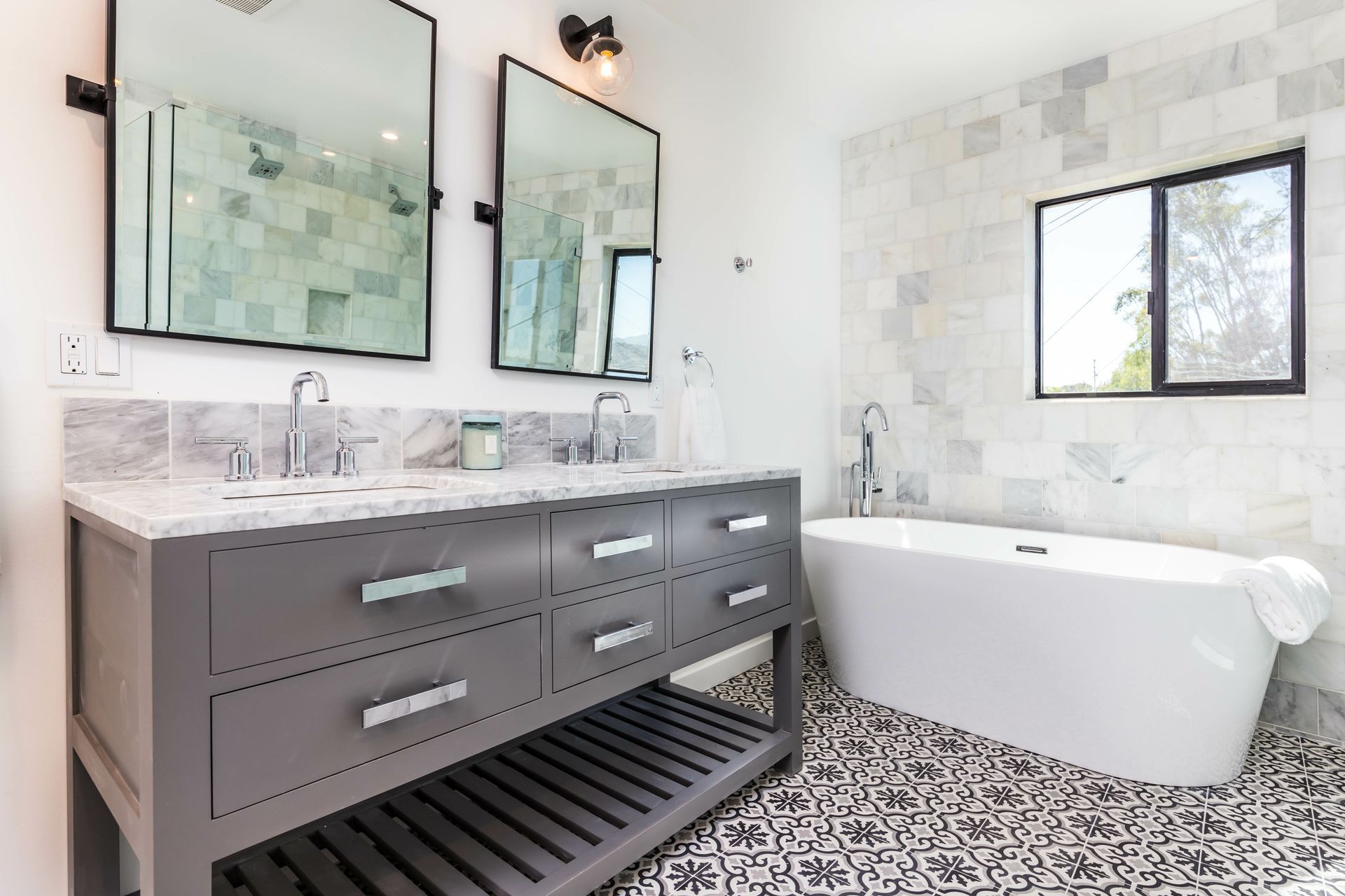 Modern bathroom with gray vanity, double mirrors, freestanding tub, and black-and-white speckled floor