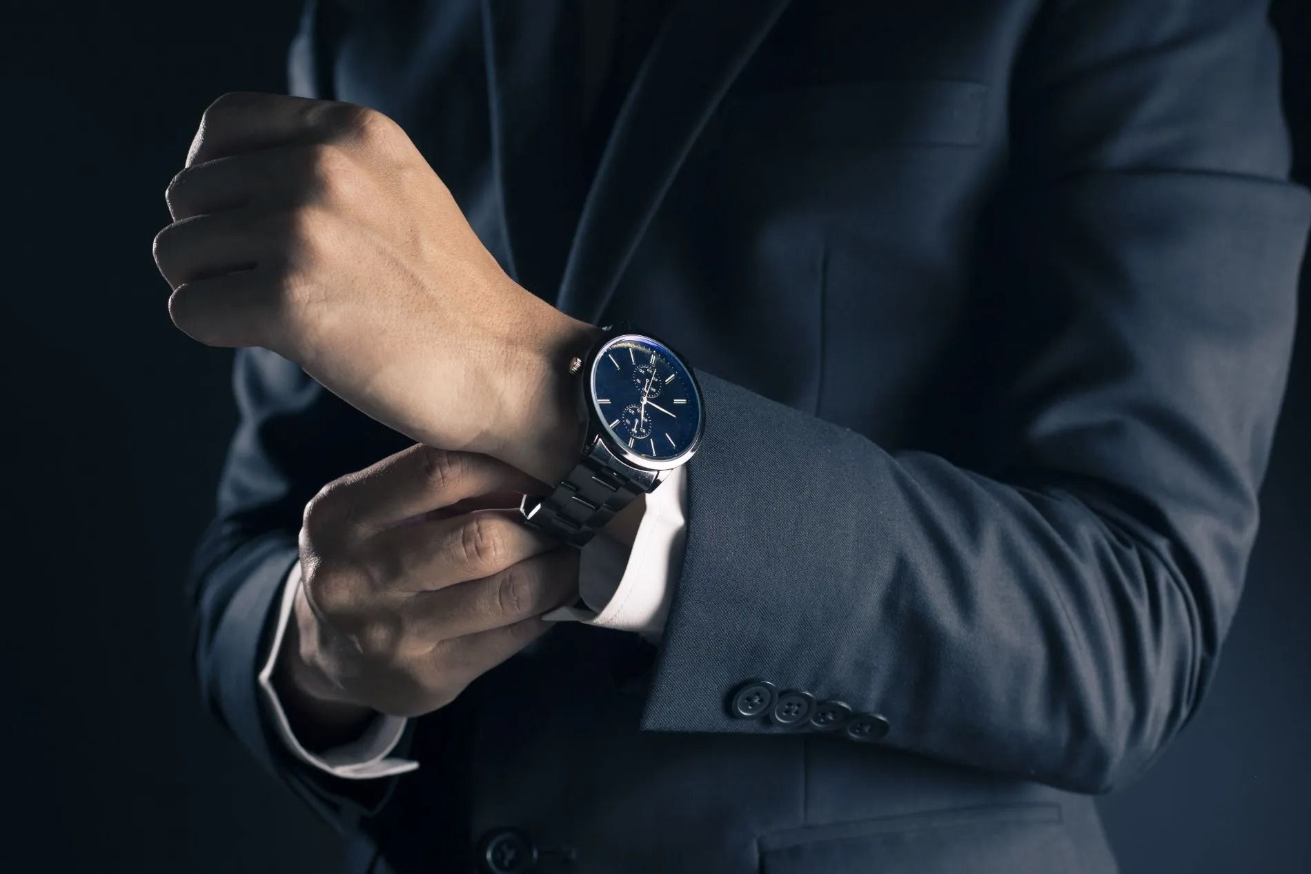 Persona che si sistema il polsino di un abito scuro, con al polso un orologio con quadrante blu, su sfondo scuro.