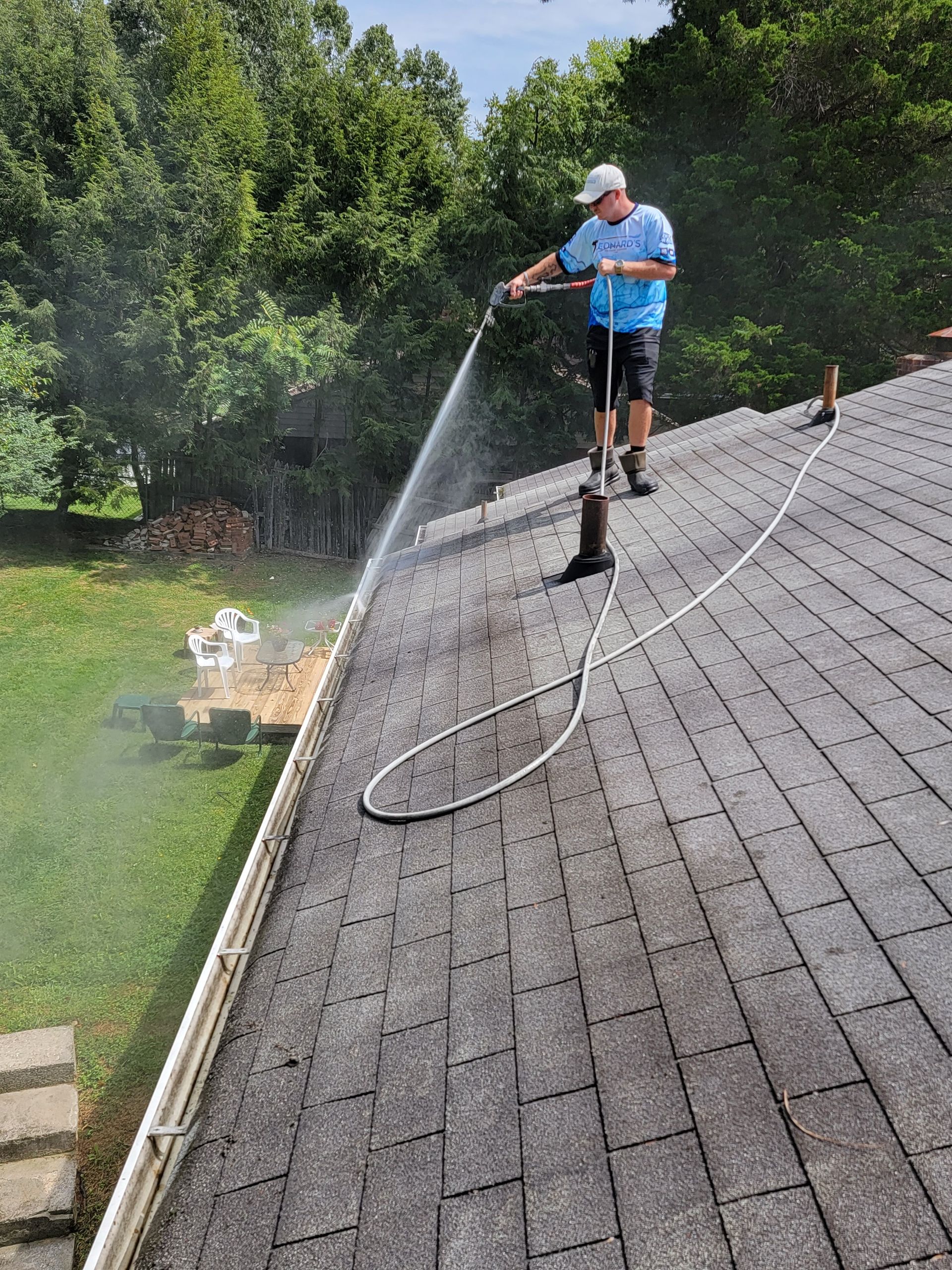Man pressure washing a dark shingle roof, spraying water. Outdoors, green trees.