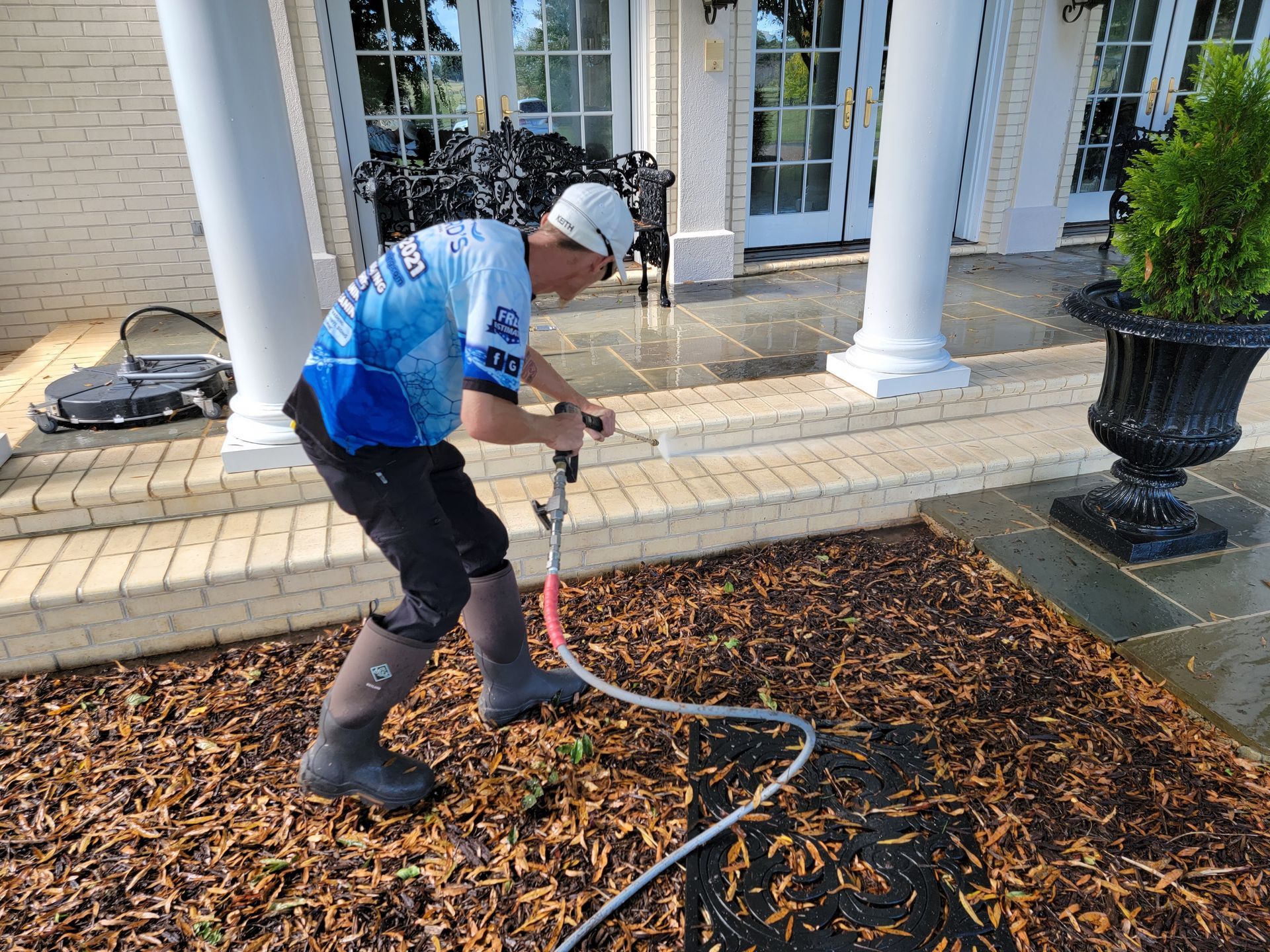 Man power washing mulch near a house with white pillars and a door, using a hose.