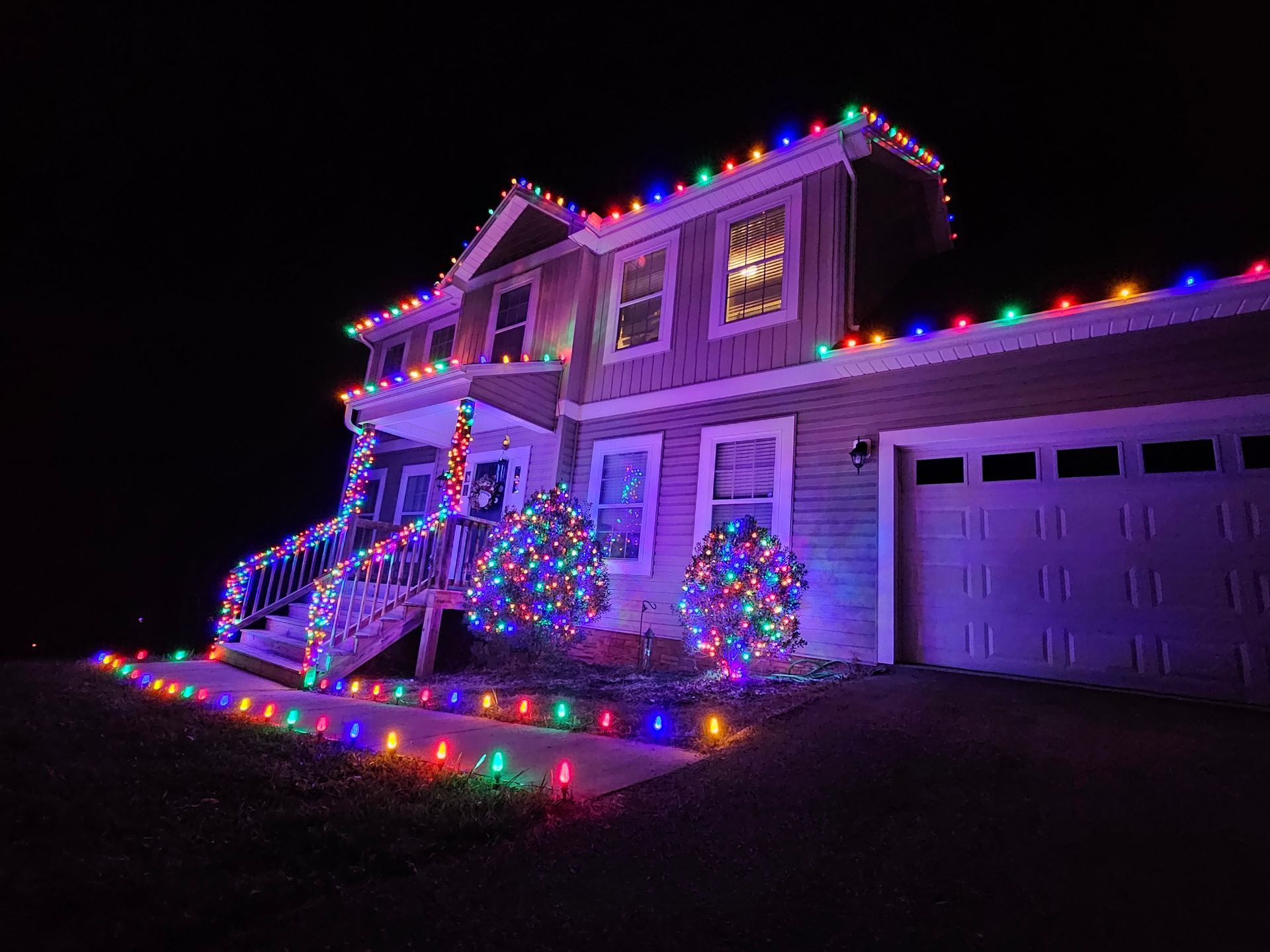 House at night, decorated with colorful Christmas lights along the roof, porch, shrubs, and walkway.
