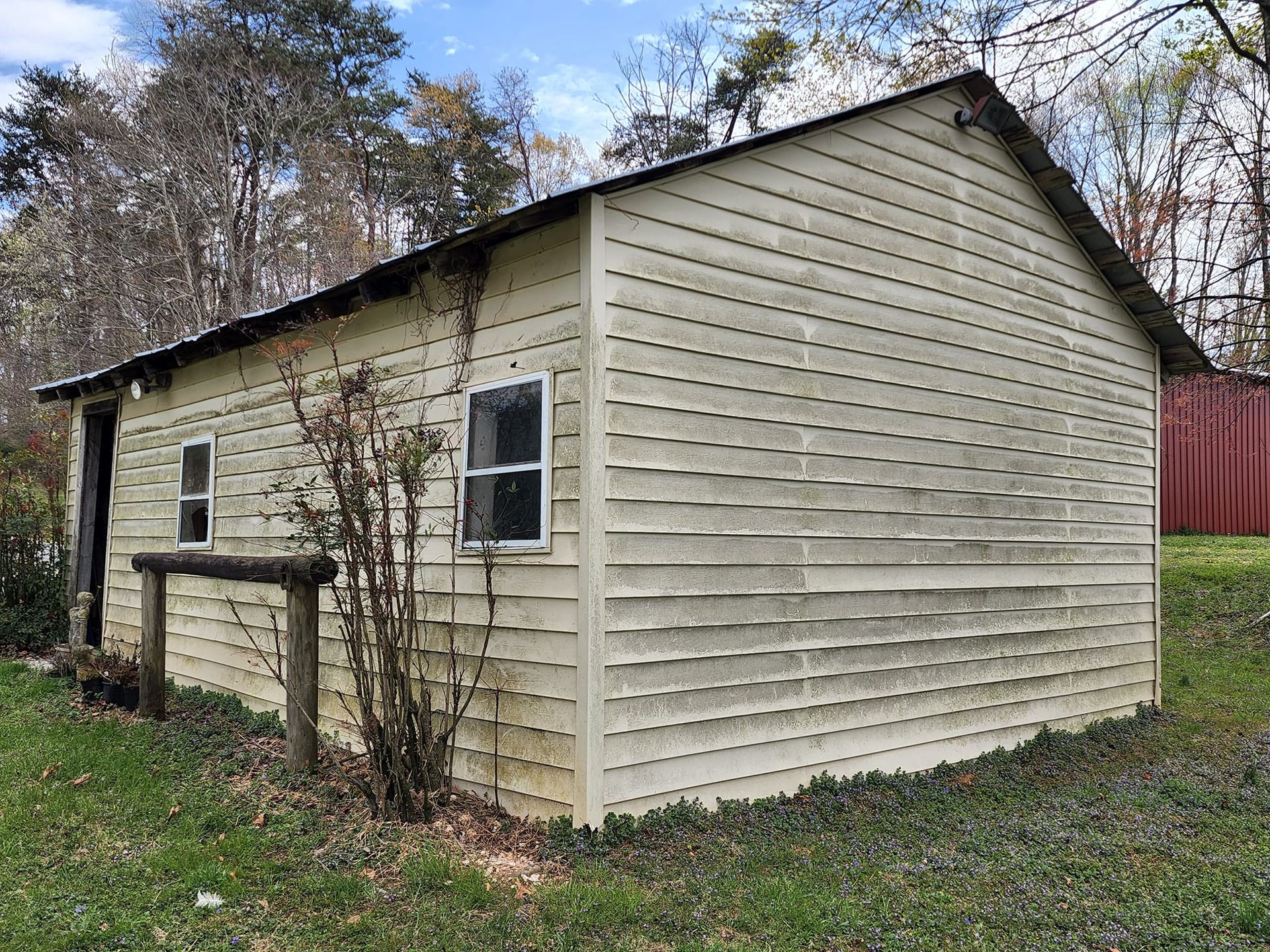 Tan, weathered shed with two windows and a metal roof, set in grassy yard near trees.