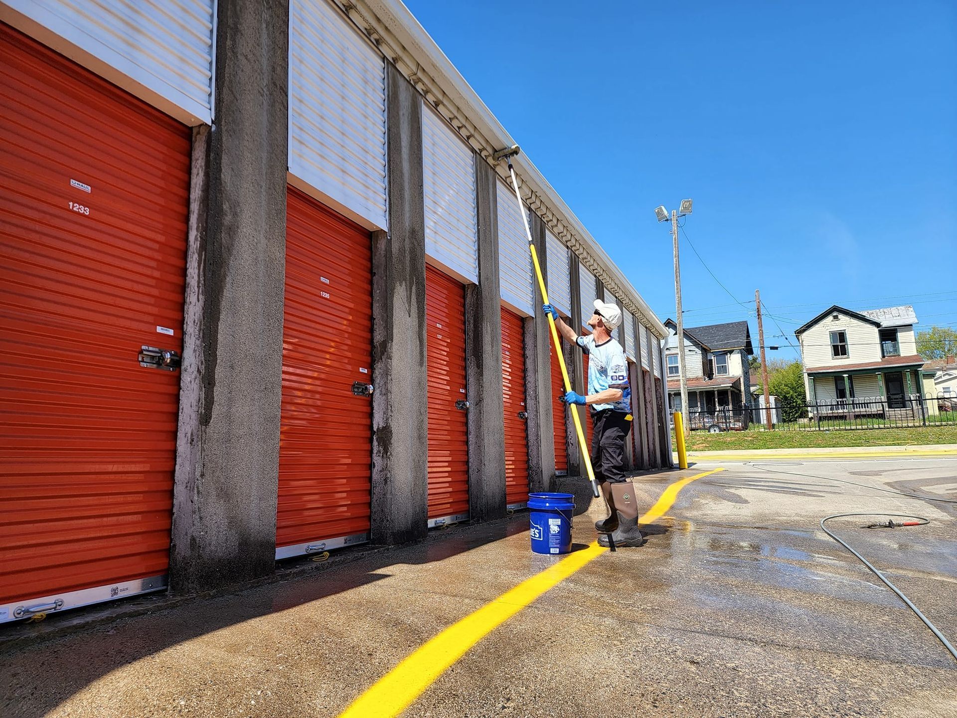 Man pressure washing storage units with red doors, outdoors on a sunny day.