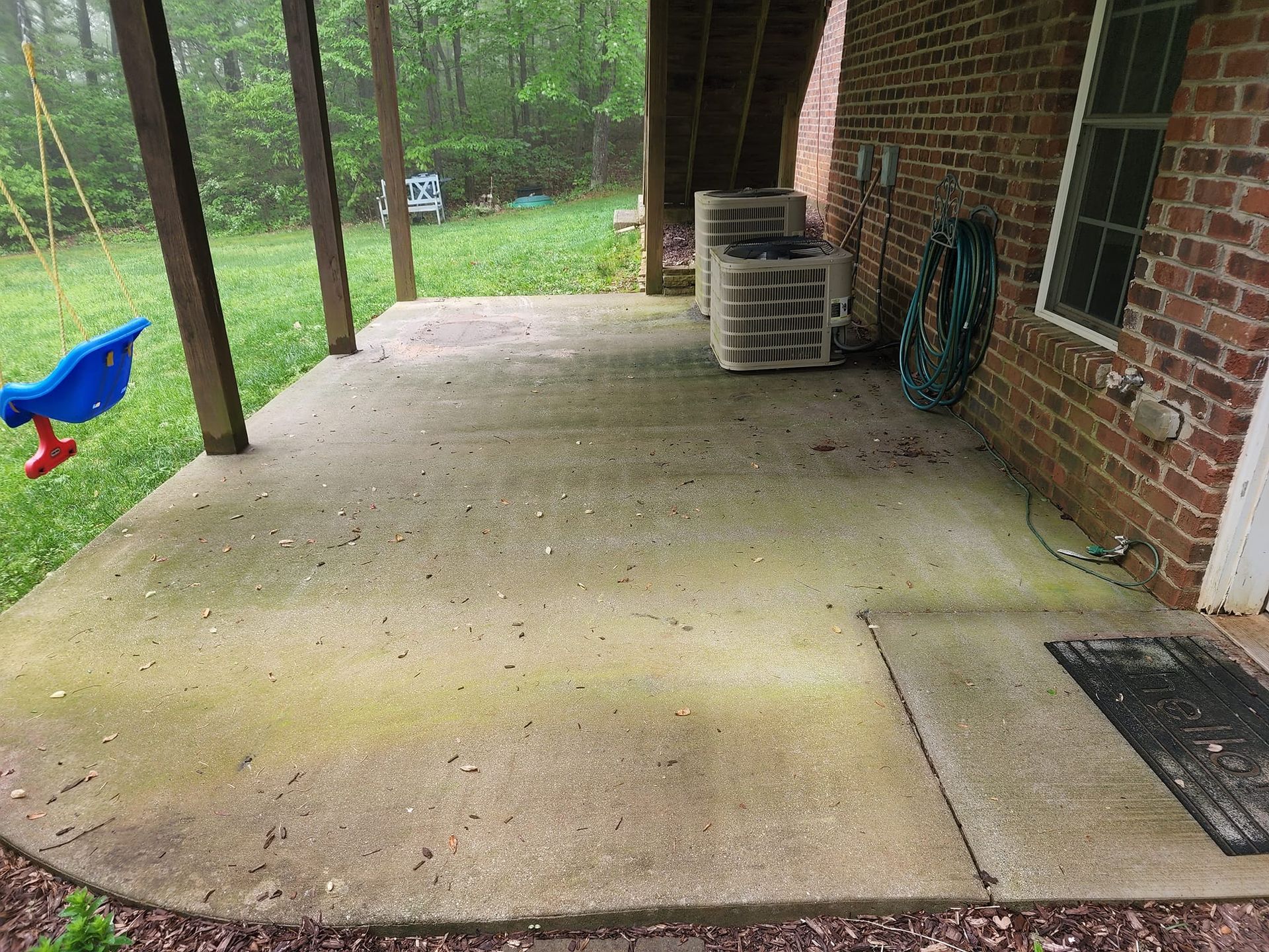 Covered concrete patio with brick wall, air conditioning units, hose, and swing set.