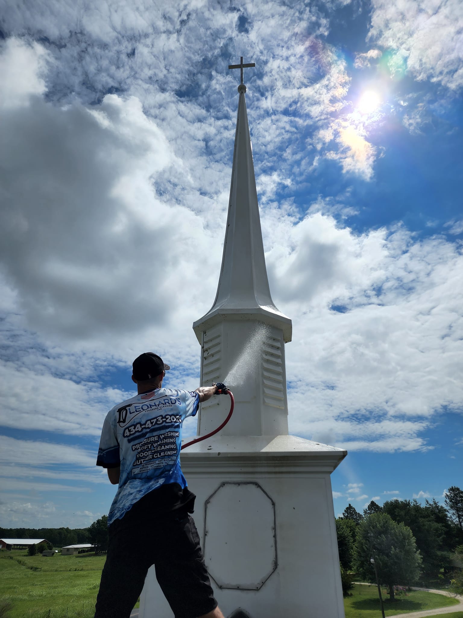 Man pressure washing a white church steeple against a bright blue sky with fluffy clouds.