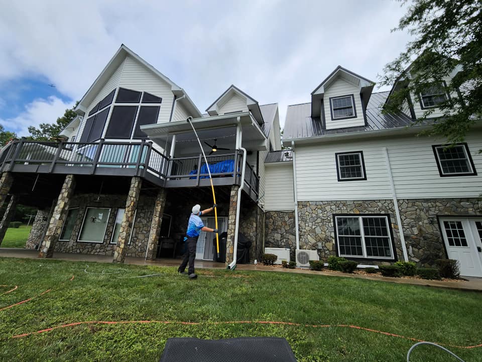 Person cleaning gutters on a large white house with a black roof and stone base, outdoors on a cloudy day.
