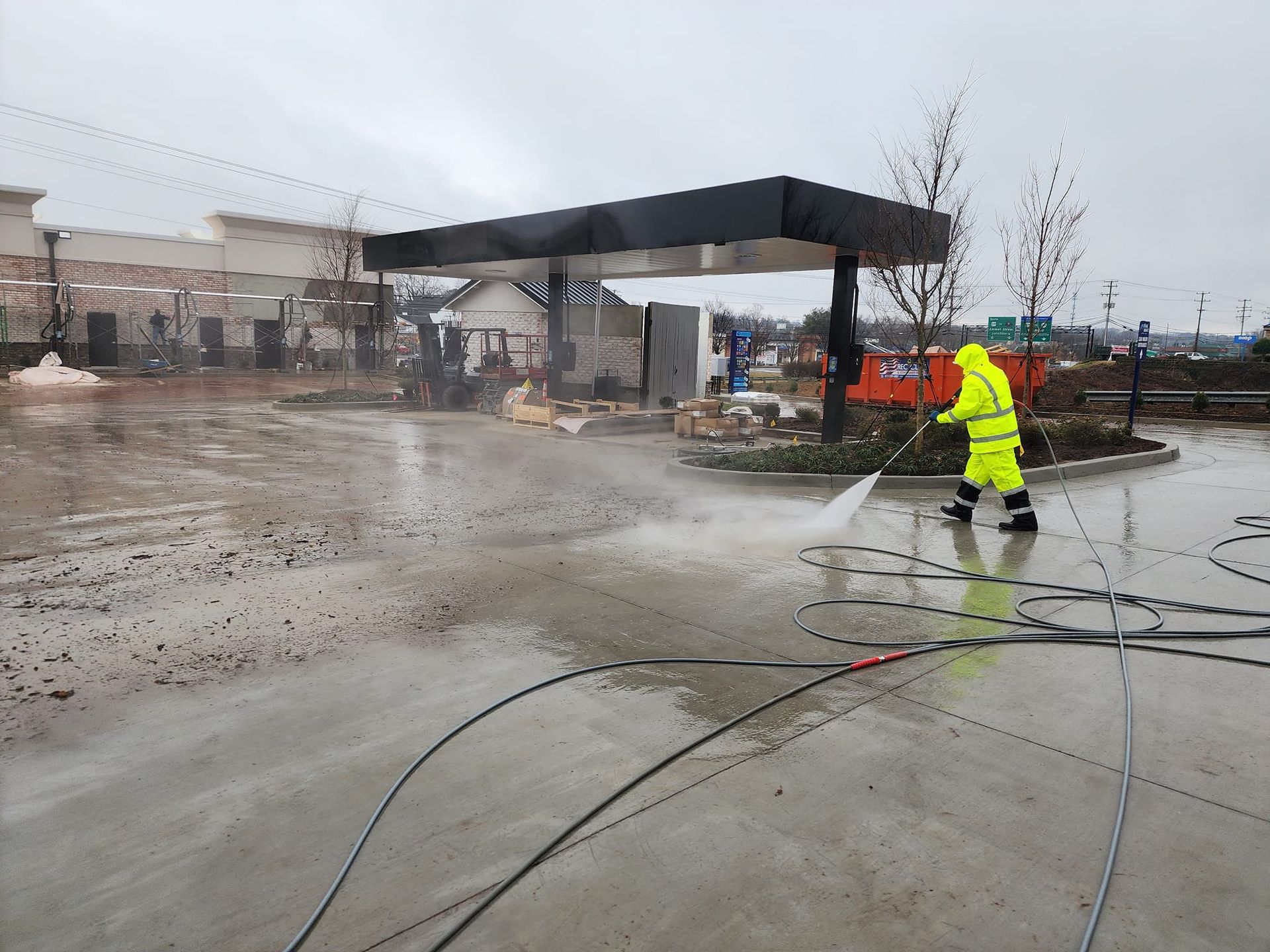 Person in yellow suit power washing a concrete area near a gas station canopy on an overcast day.