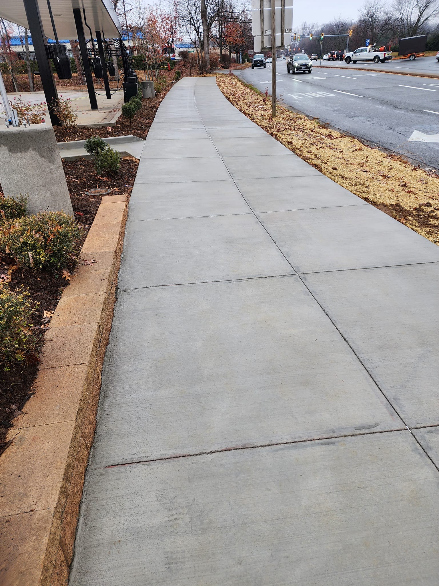 Concrete sidewalk next to a road, edged by a retaining wall and landscaping.