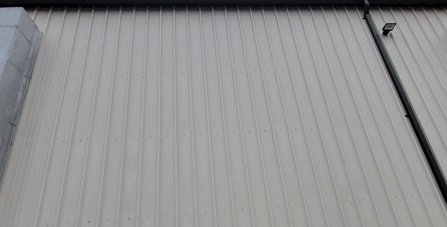 Beige metal siding on a building exterior, vertical grooves, with a black pipe and small light.