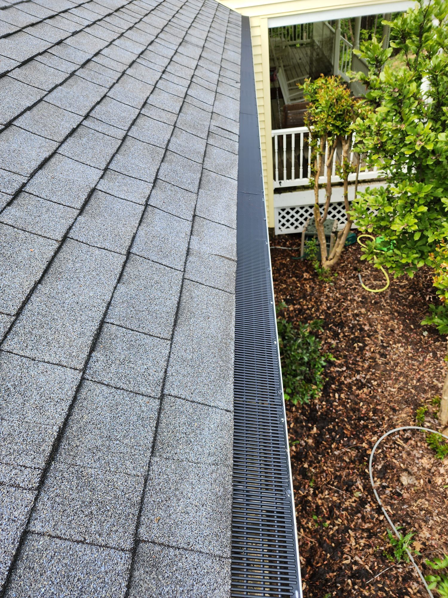Roof with gray shingles and black gutter, next to a yard with mulch and a house.