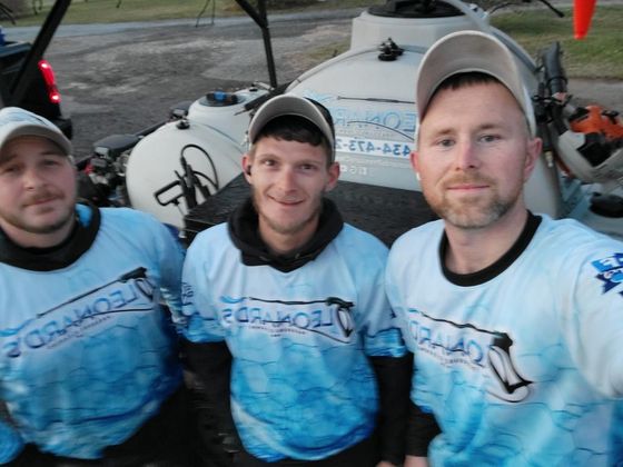 Three men in matching blue shirts with logo stand in front of a tank and equipment.