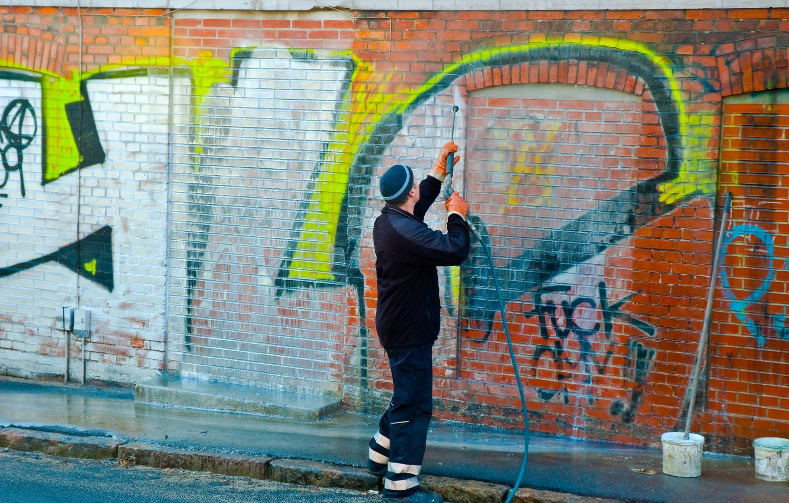 Person cleaning graffiti from a brick wall, using water and a long handled brush.