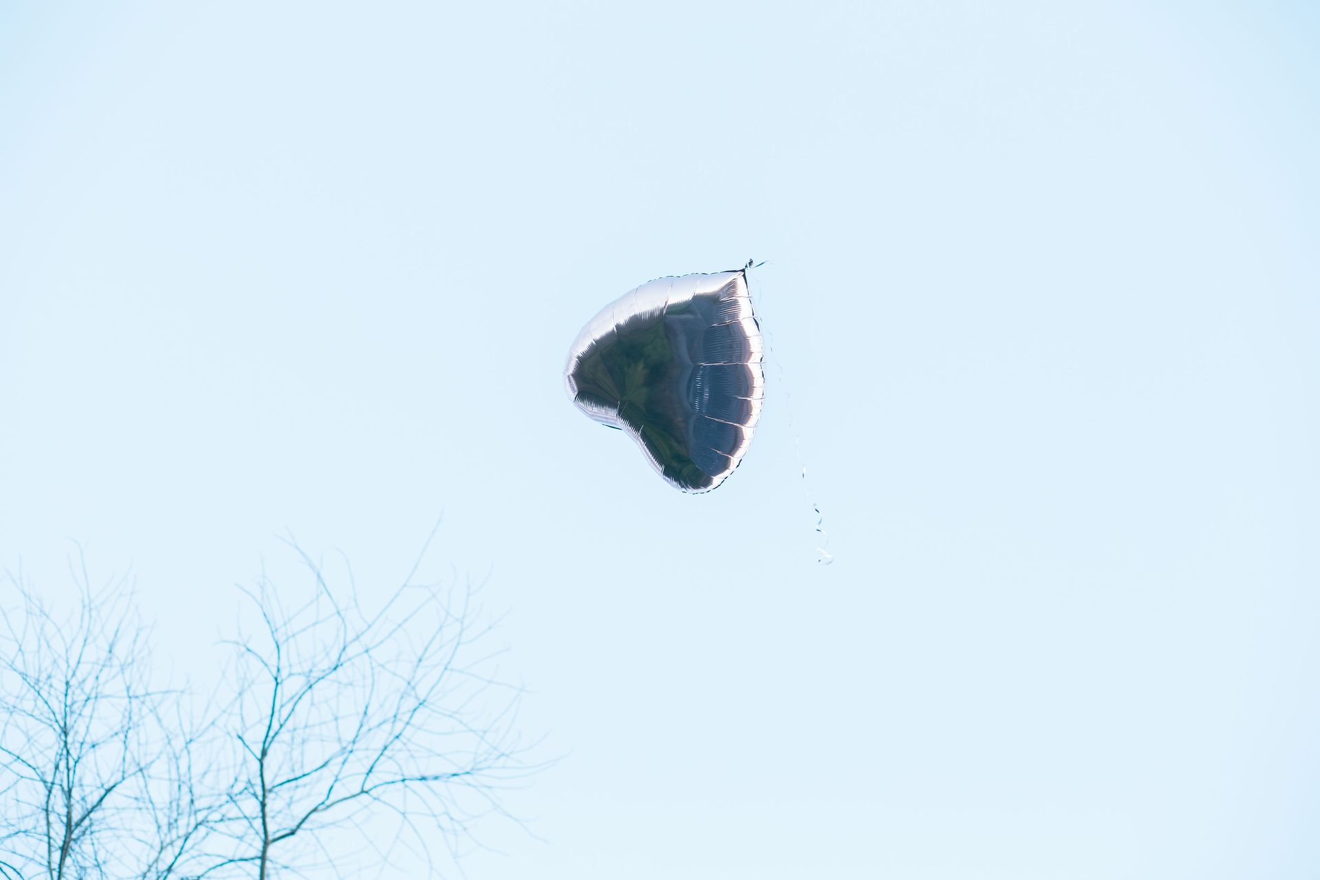 A dark, deflated balloon floating in a clear blue sky with bare tree branches