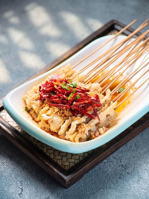 Grilled meat skewers on a white dish, topped with red chili and herbs, on a dark tray.