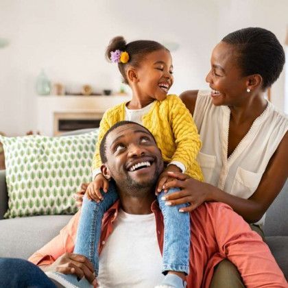 A man and woman are sitting on a couch with a little girl on his shoulders.
