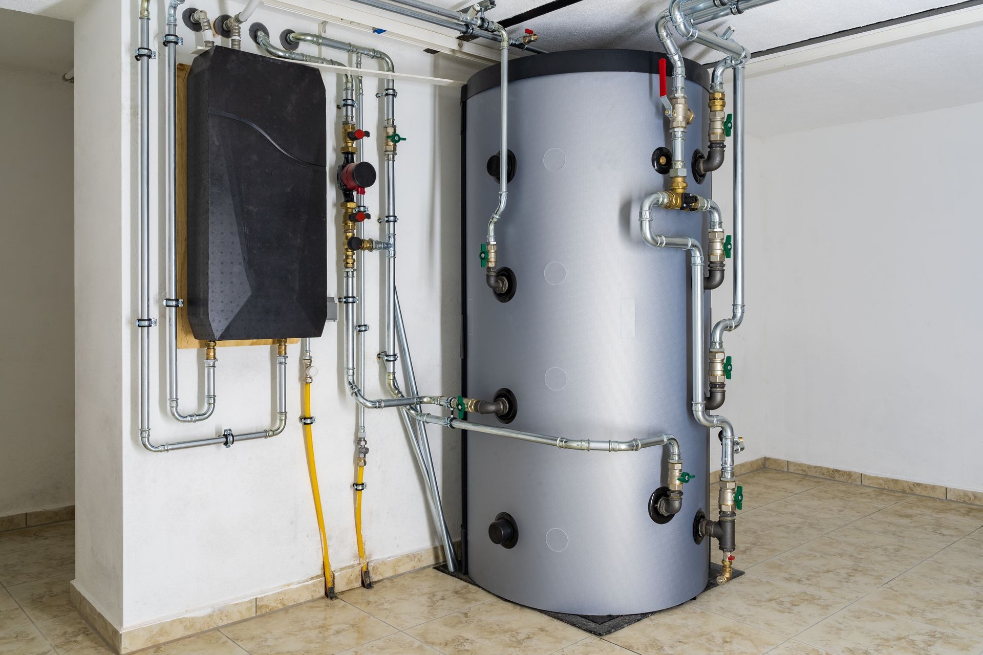 Large gray water heater and black rectangular heat pump with connecting pipes in a white-walled room.