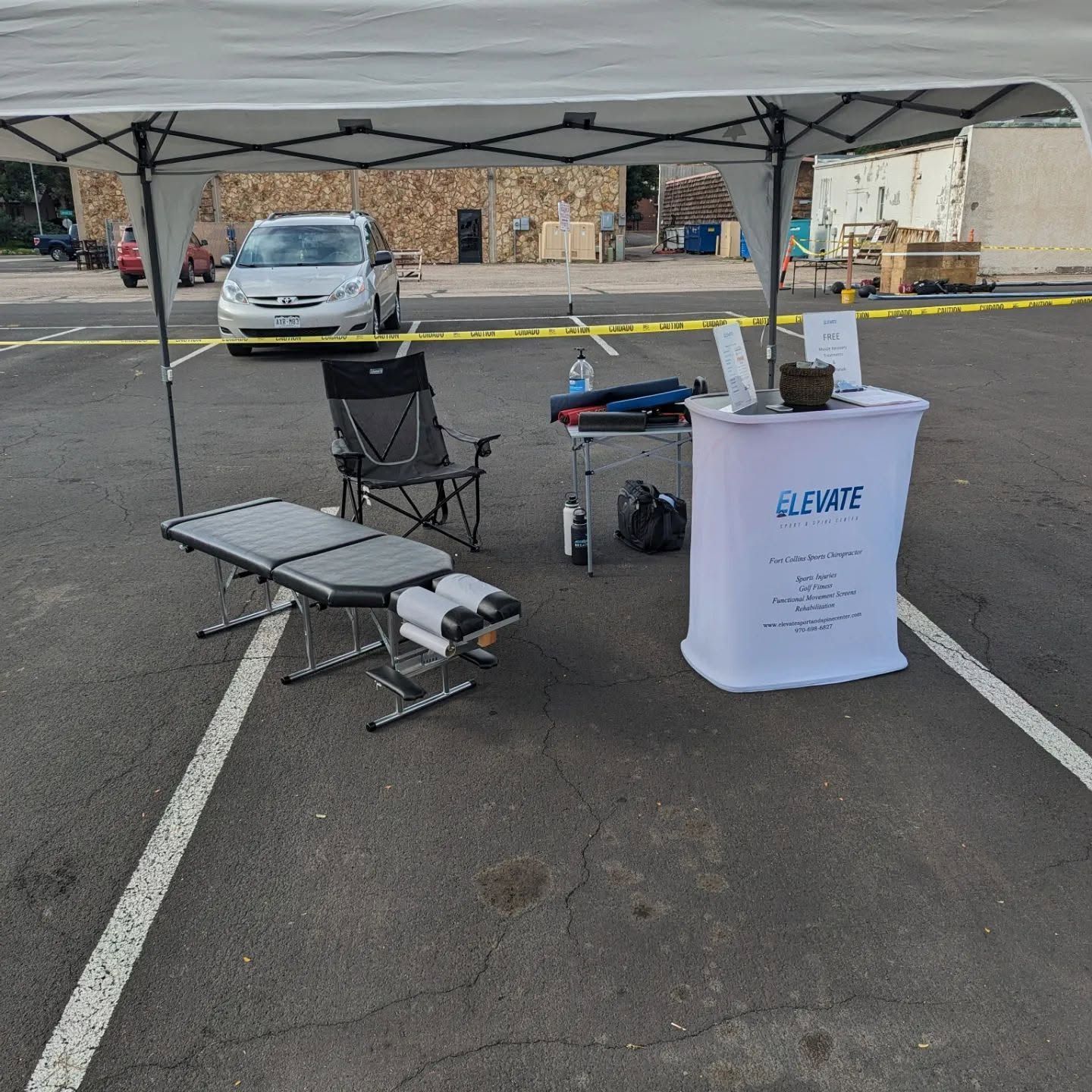 Under a white canopy, a foldable table and chair sit beside a branded table in a parking lot