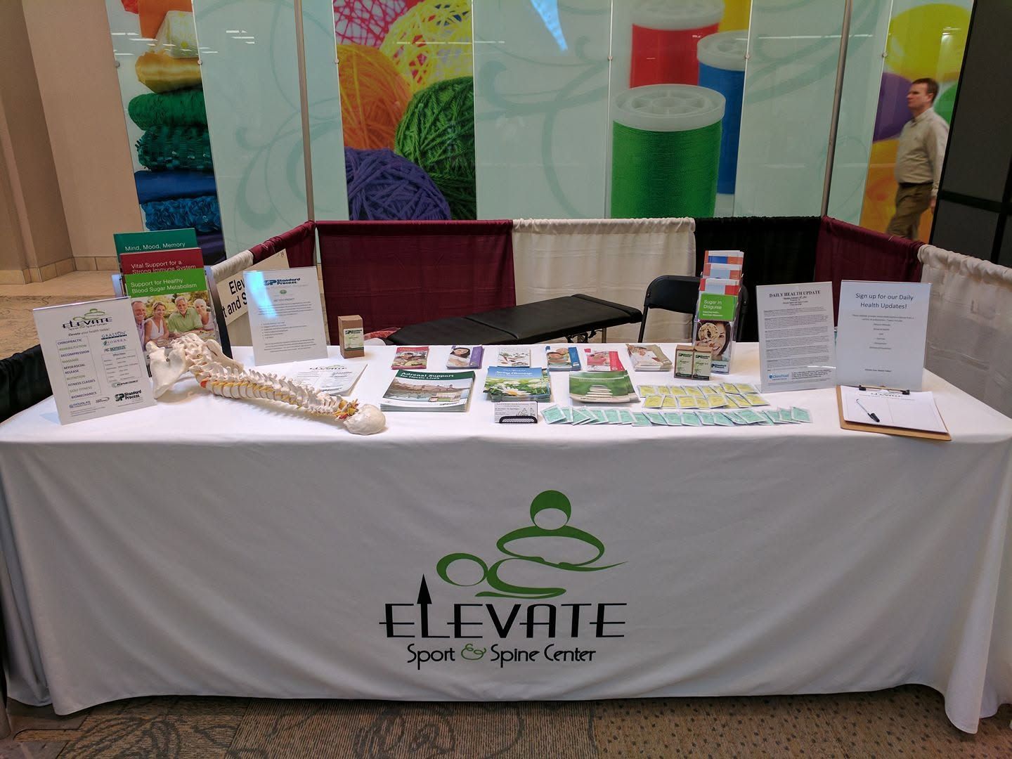 Vendor booth with white tablecloth and green logo, displaying small packaged goods and signs at a fair.