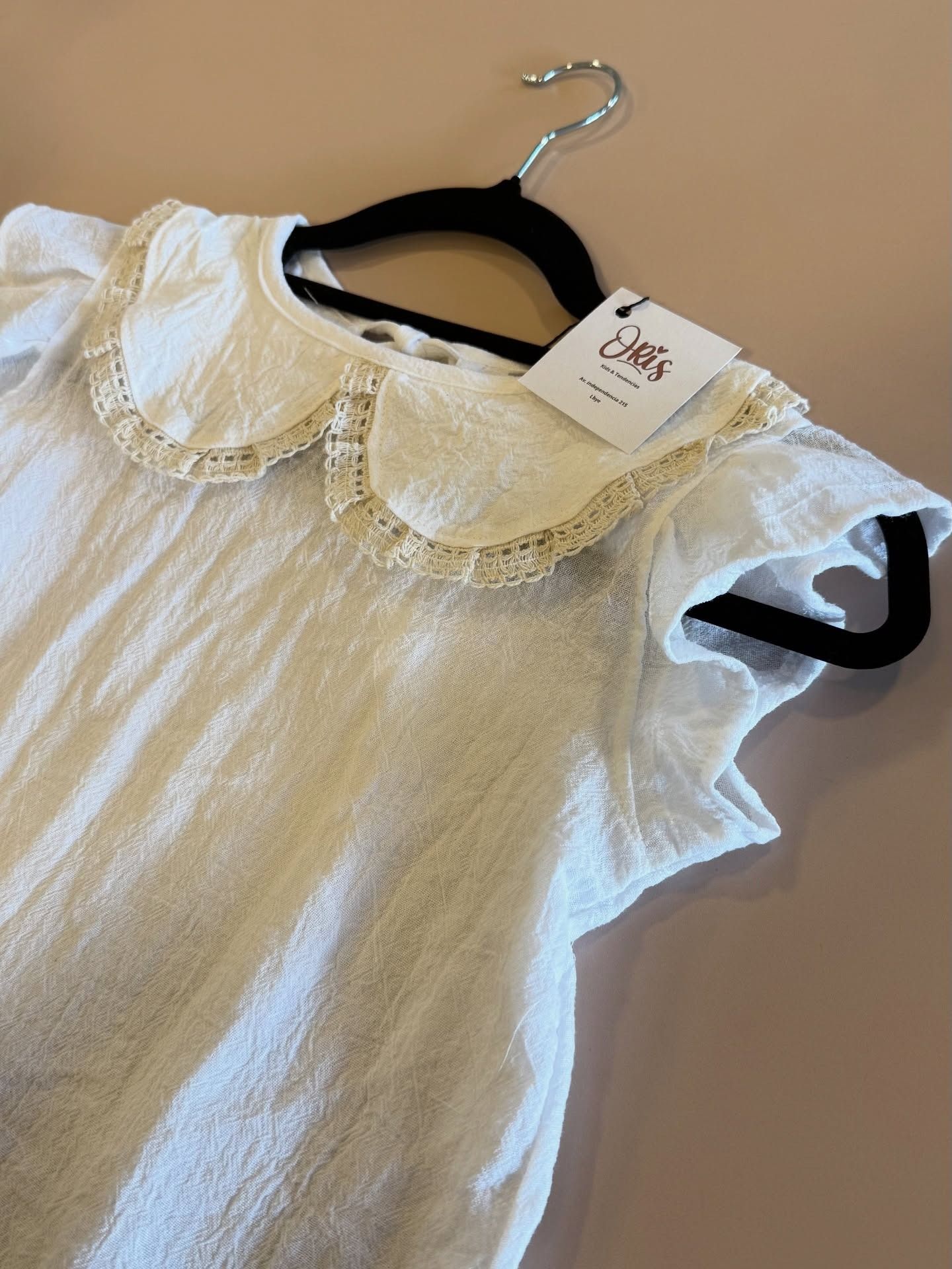 Blusa blanca con cuello festoneado en una percha negra.