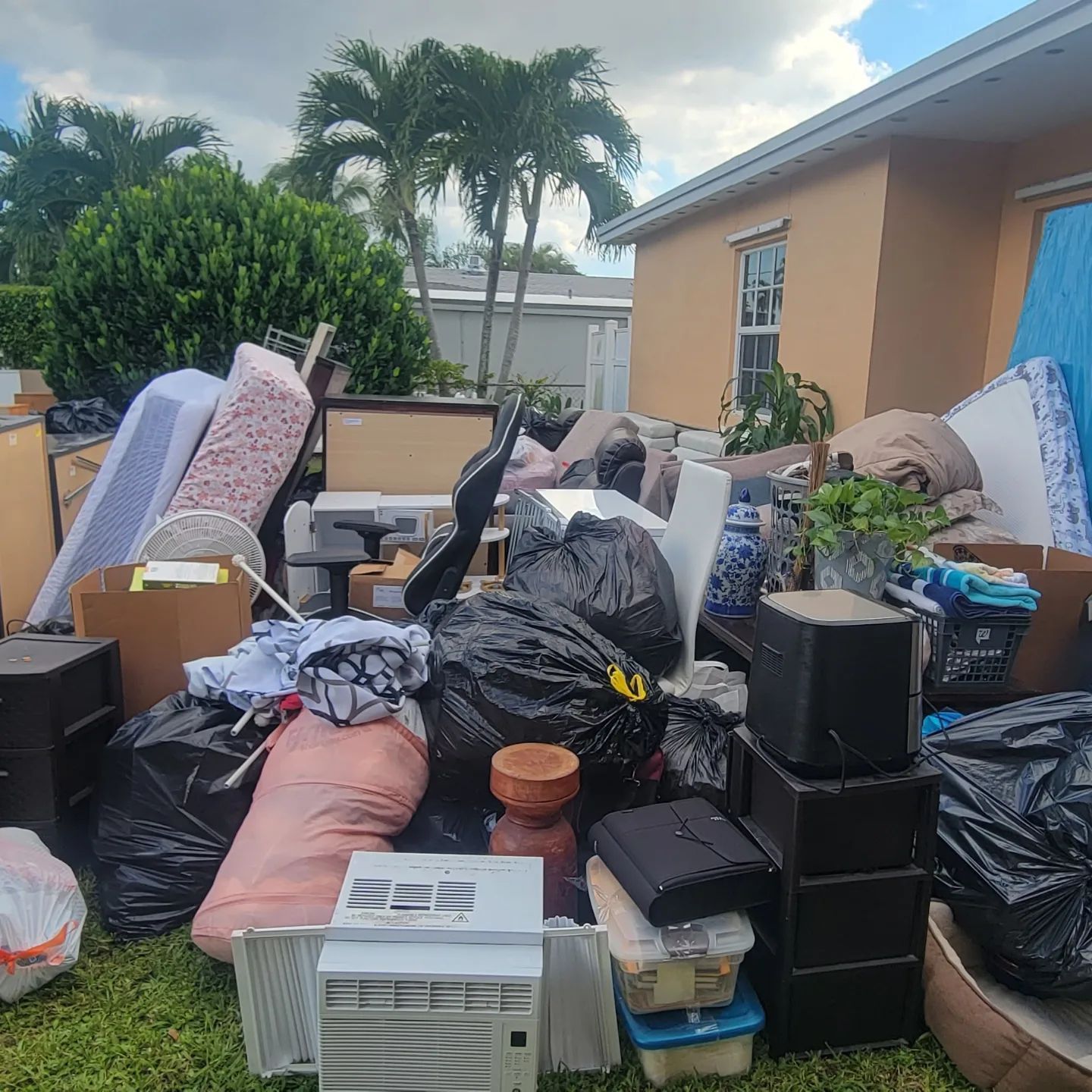 Pile of household junk and trash in a yard, including furniture, bags, and an air conditioner.