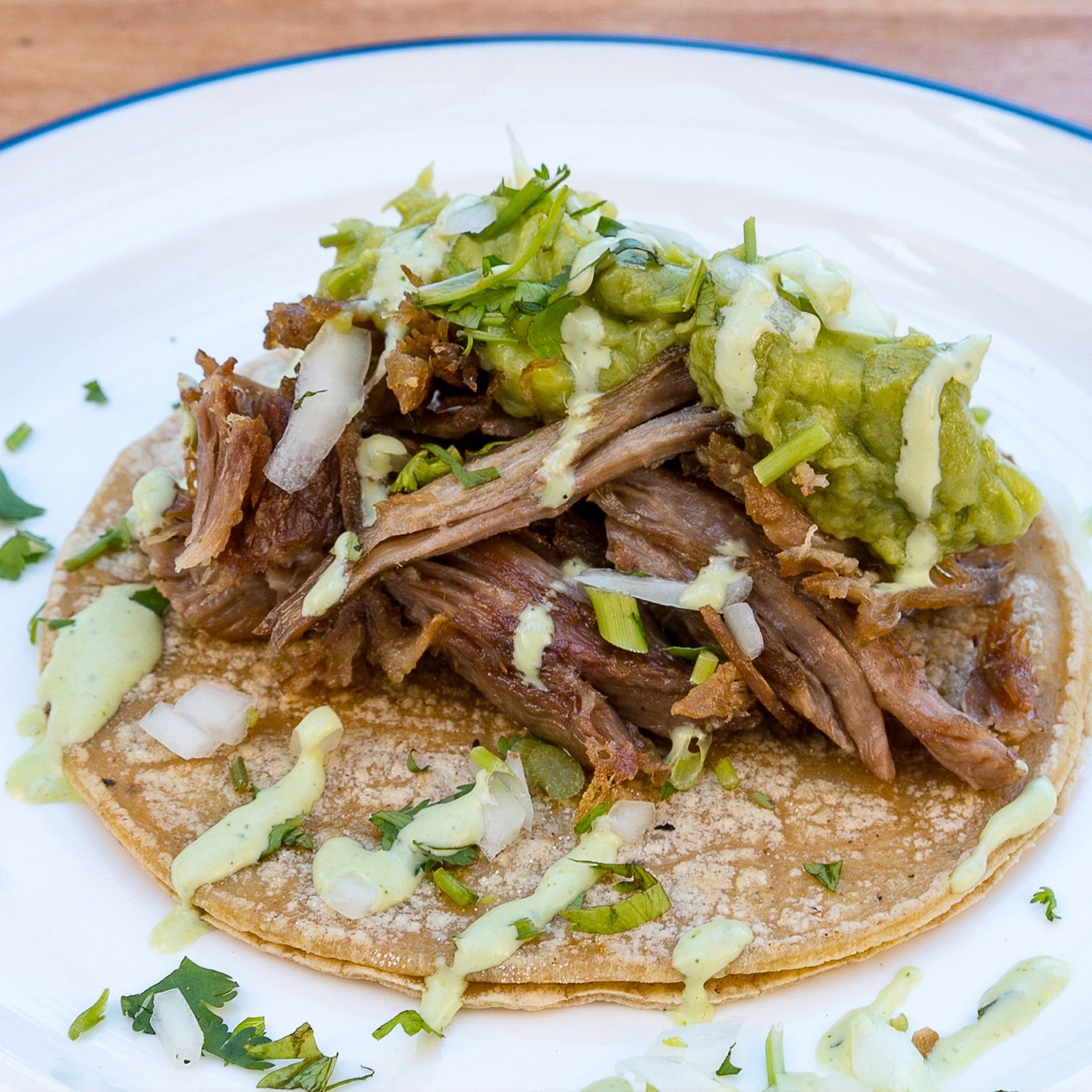 Taco on a white plate, topped with shredded meat, guacamole, onions, cilantro, and green sauce.