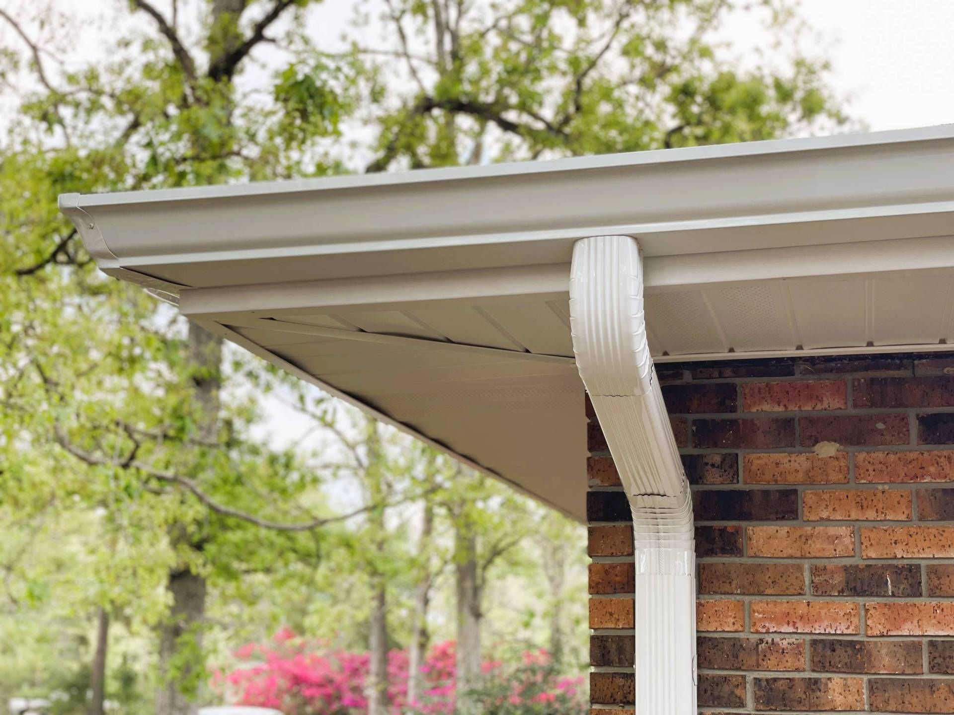 White gutter and downspout on a brick house roofline, with trees and pink flowers in the background