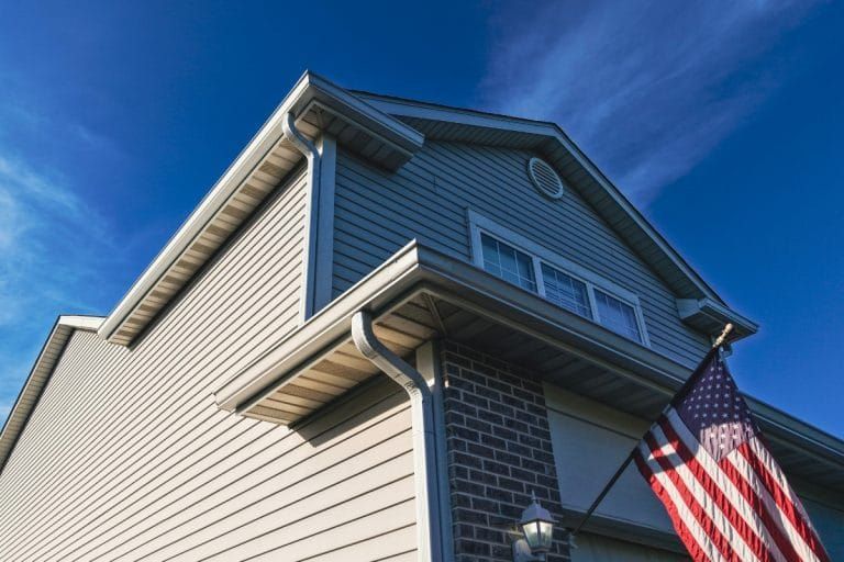 Low-angle view of a house with beige siding, brick accents, and an American flag against a blue sky