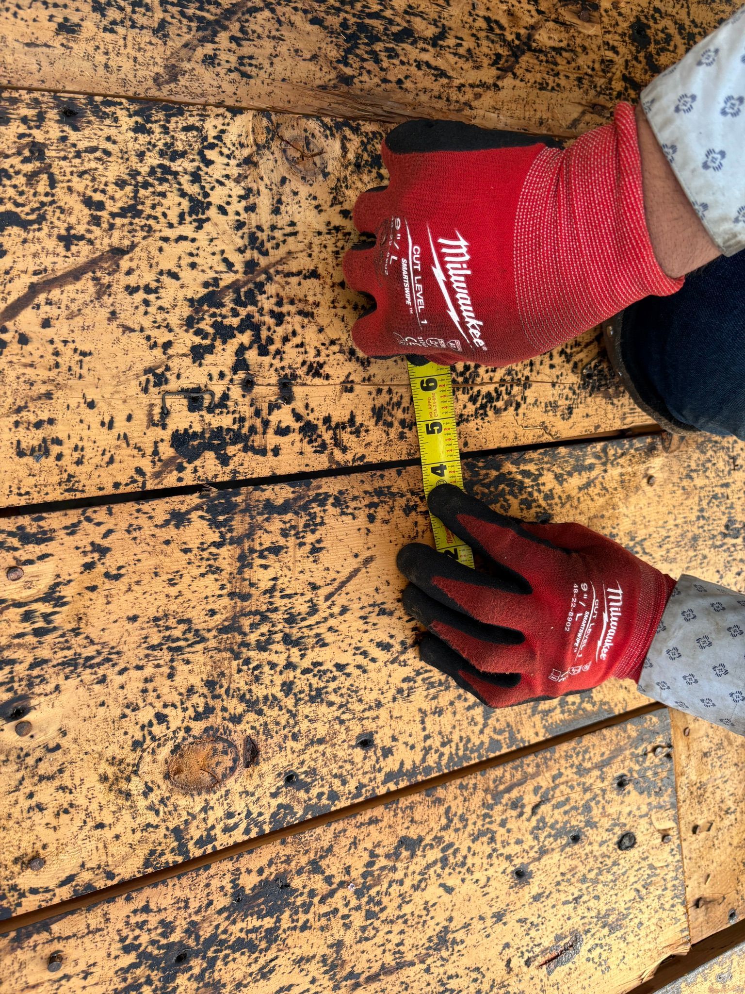 Gloved hands measure the width of tan roof shingles with a yellow tape measure.