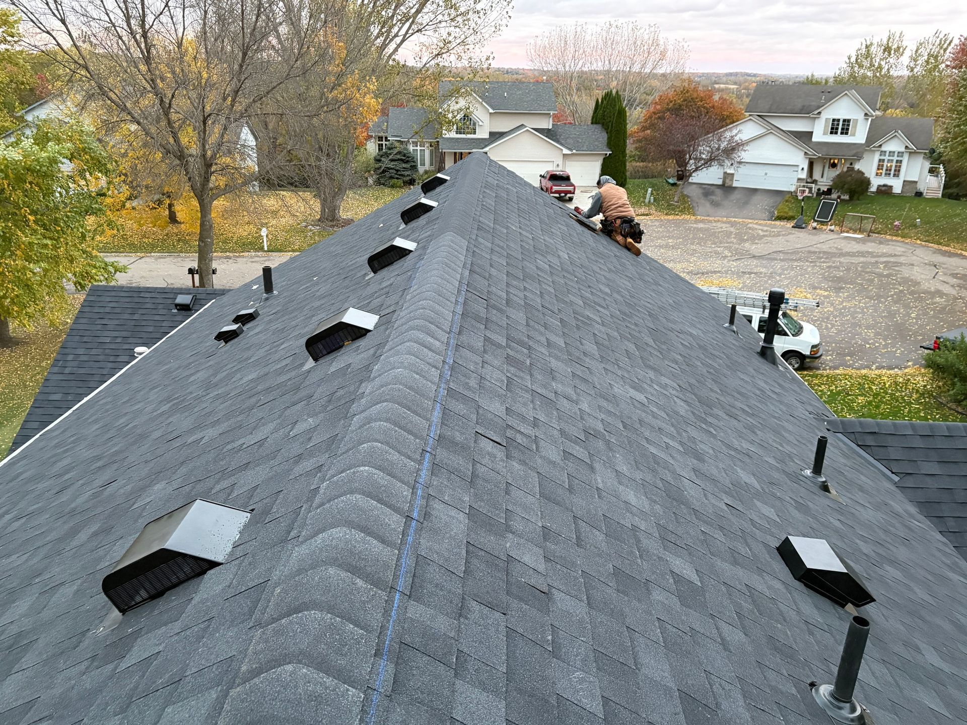 Roof under construction, viewed from the ridge, with shingles, vents, and workers in the distance.