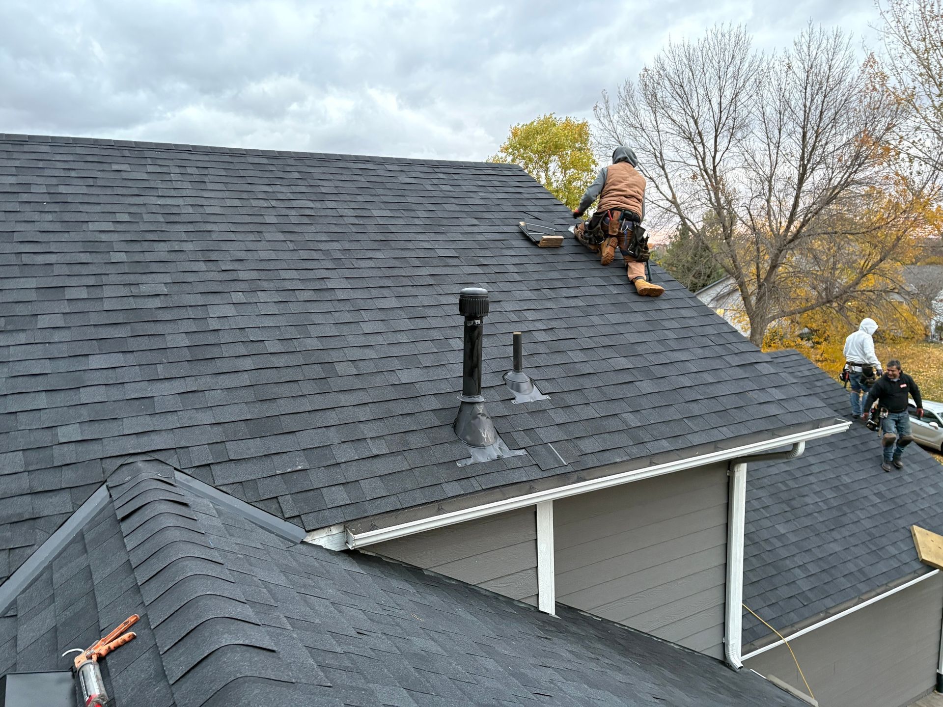 Workers repairing a gray shingle roof on a house, with tools and supplies visible.