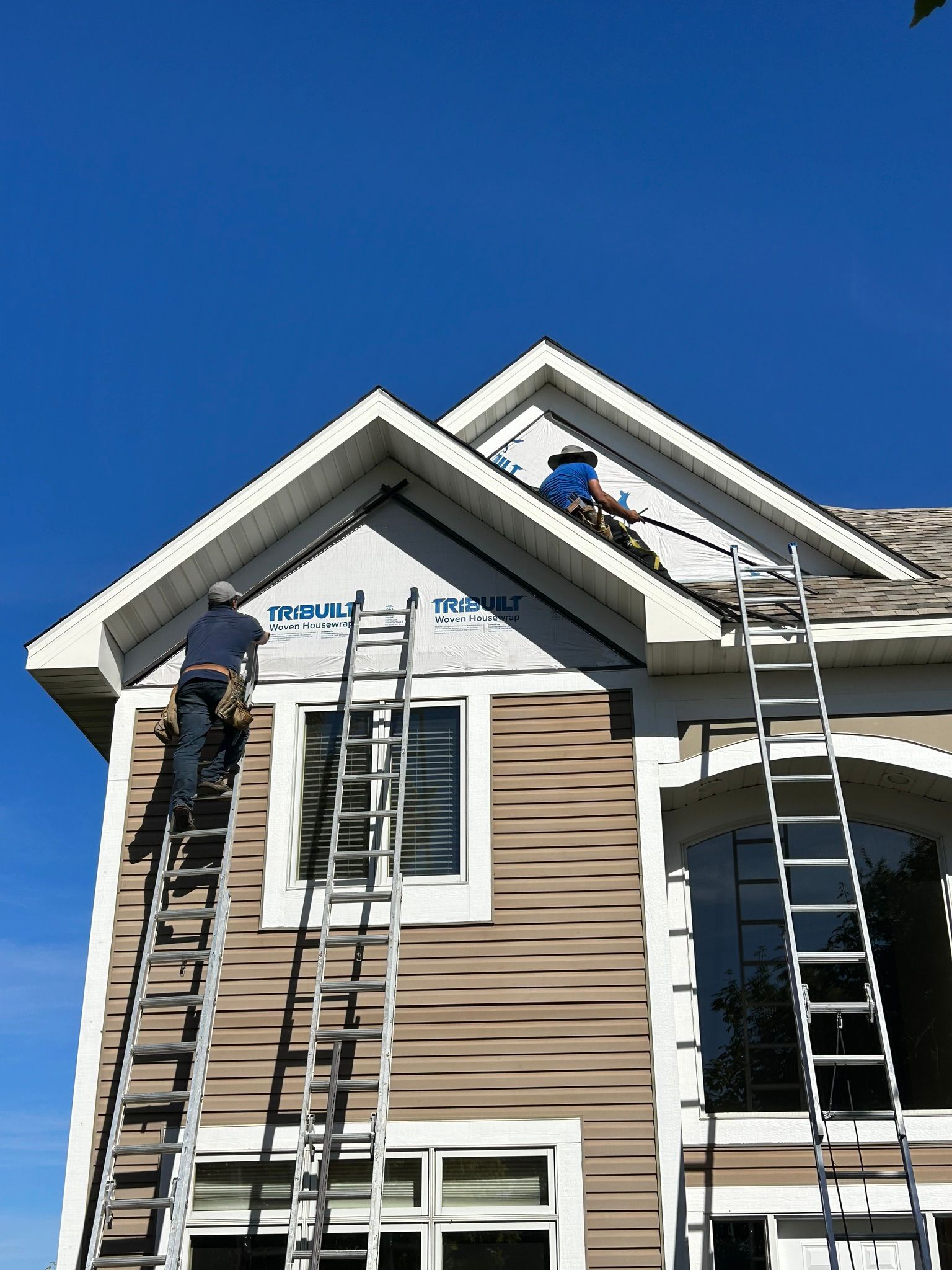 Two workers on ladders painting the front gable of a tan house under a clear blue sky