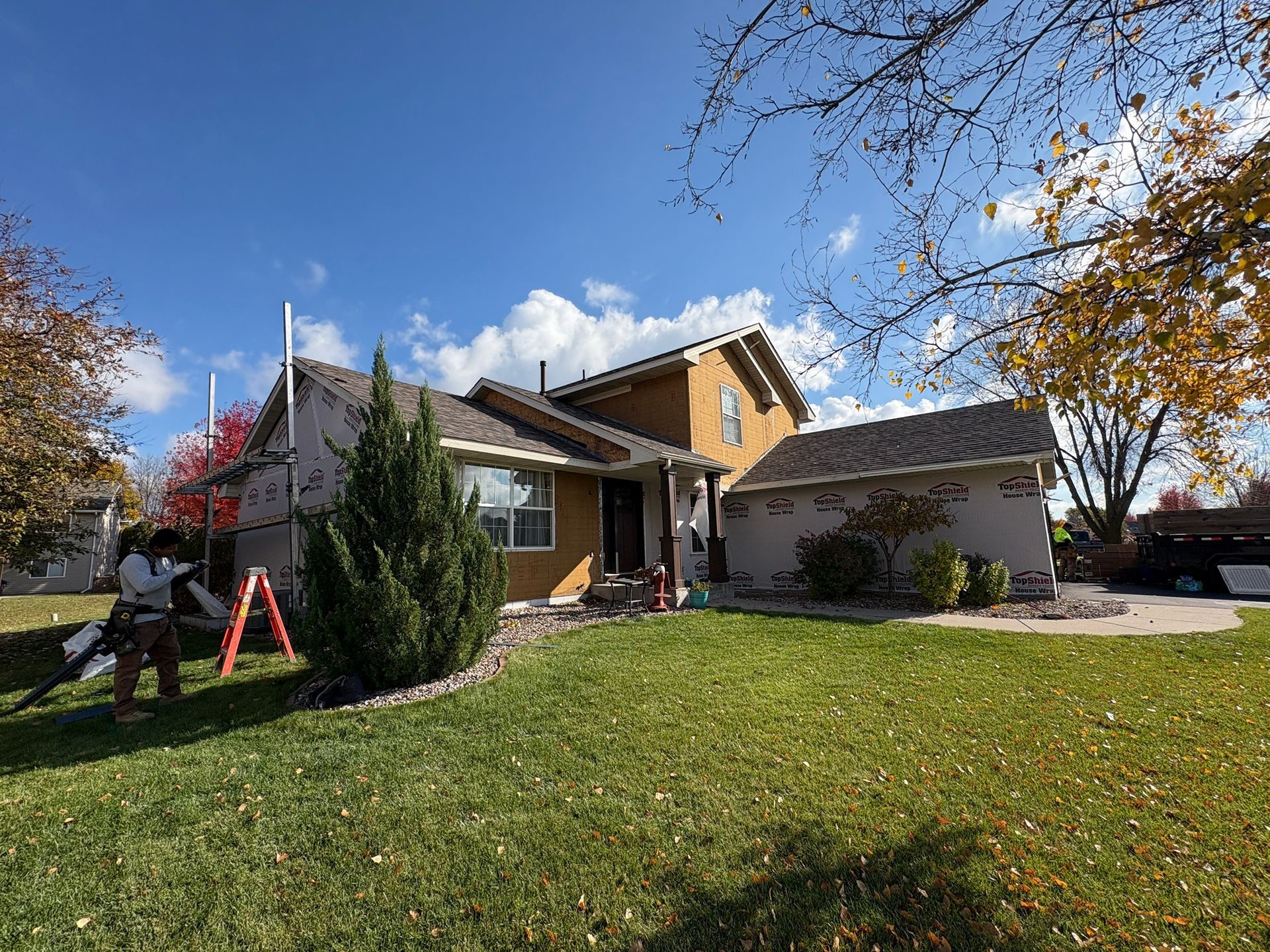 Suburban house with a green lawn, autumn trees, and a parked car under a blue sky.