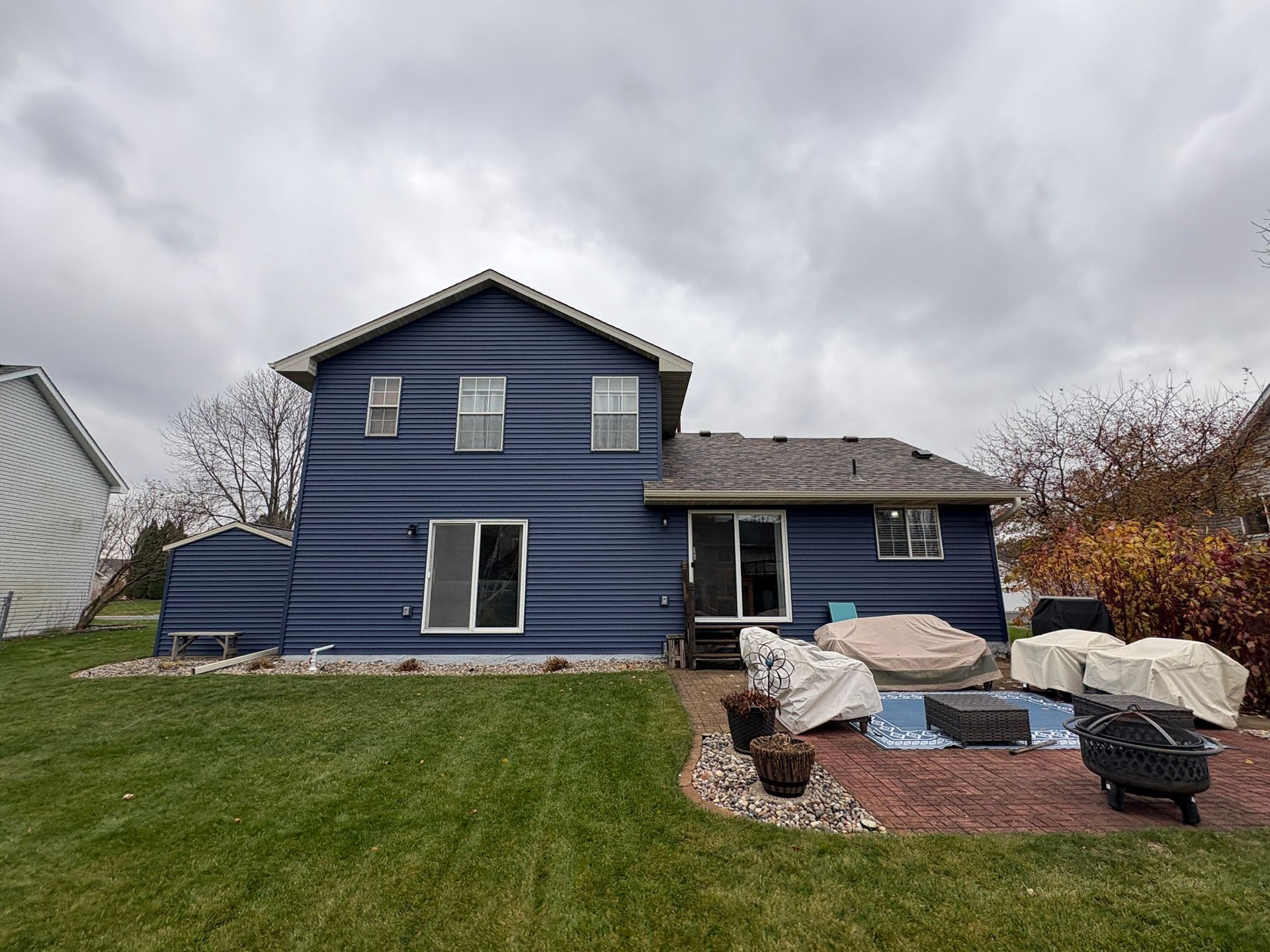 Blue two-story house with a backyard patio and covered furniture under a cloudy sky