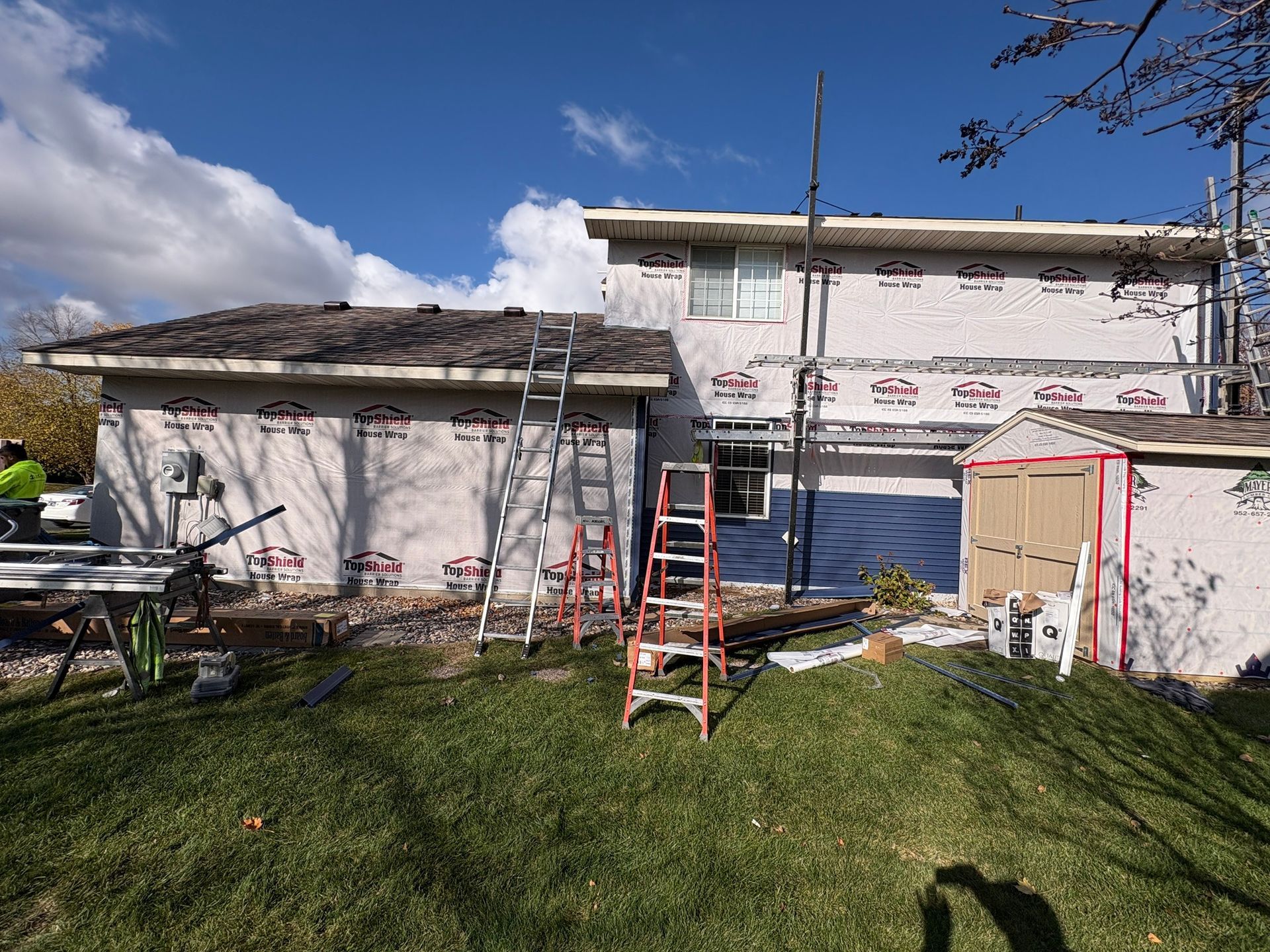 Partially renovated house with scaffolding and ladders in a grassy yard under a blue sky