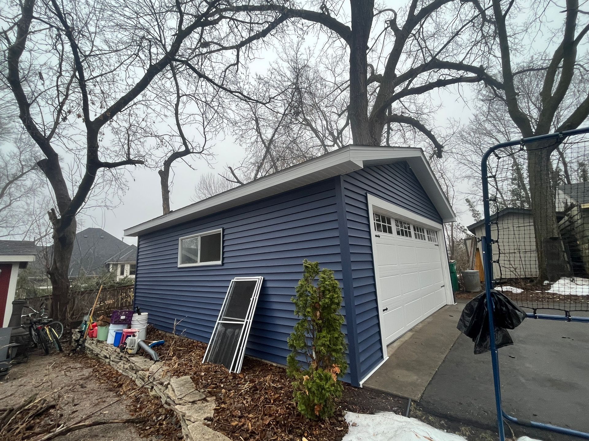 Blue detached garage with white door and ladder in a snowy yard under bare trees