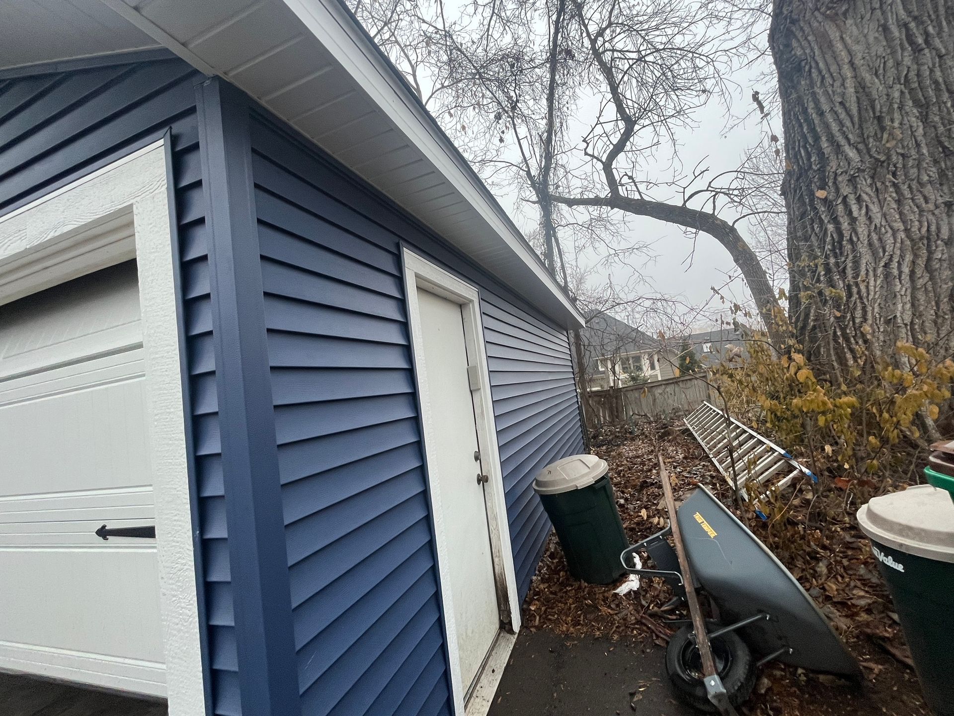 Blue-sided garage beside a tree, with a white door and fallen leaves along the side yard.