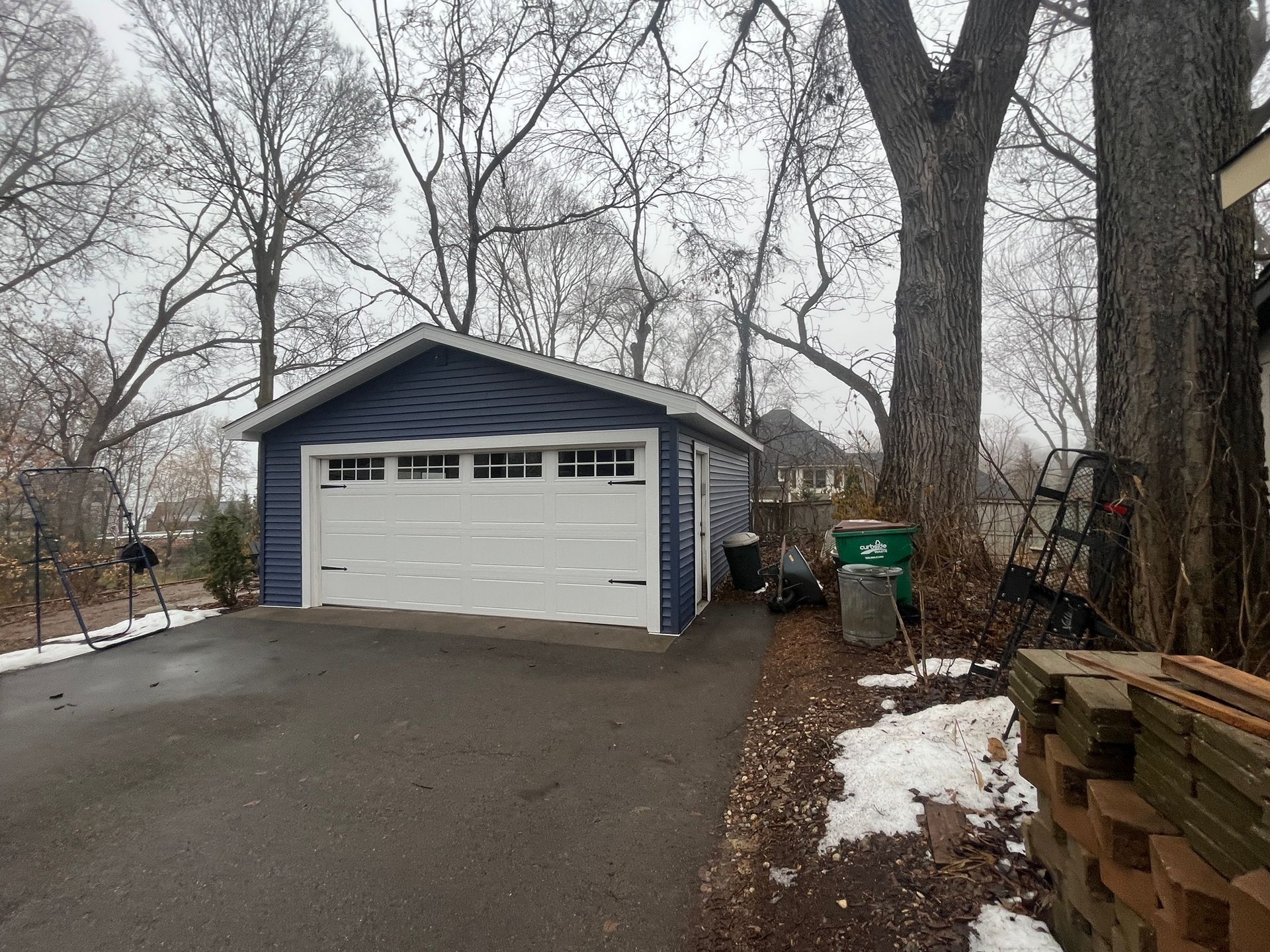 Detached blue garage with white door beside a snowy driveway and leafless trees