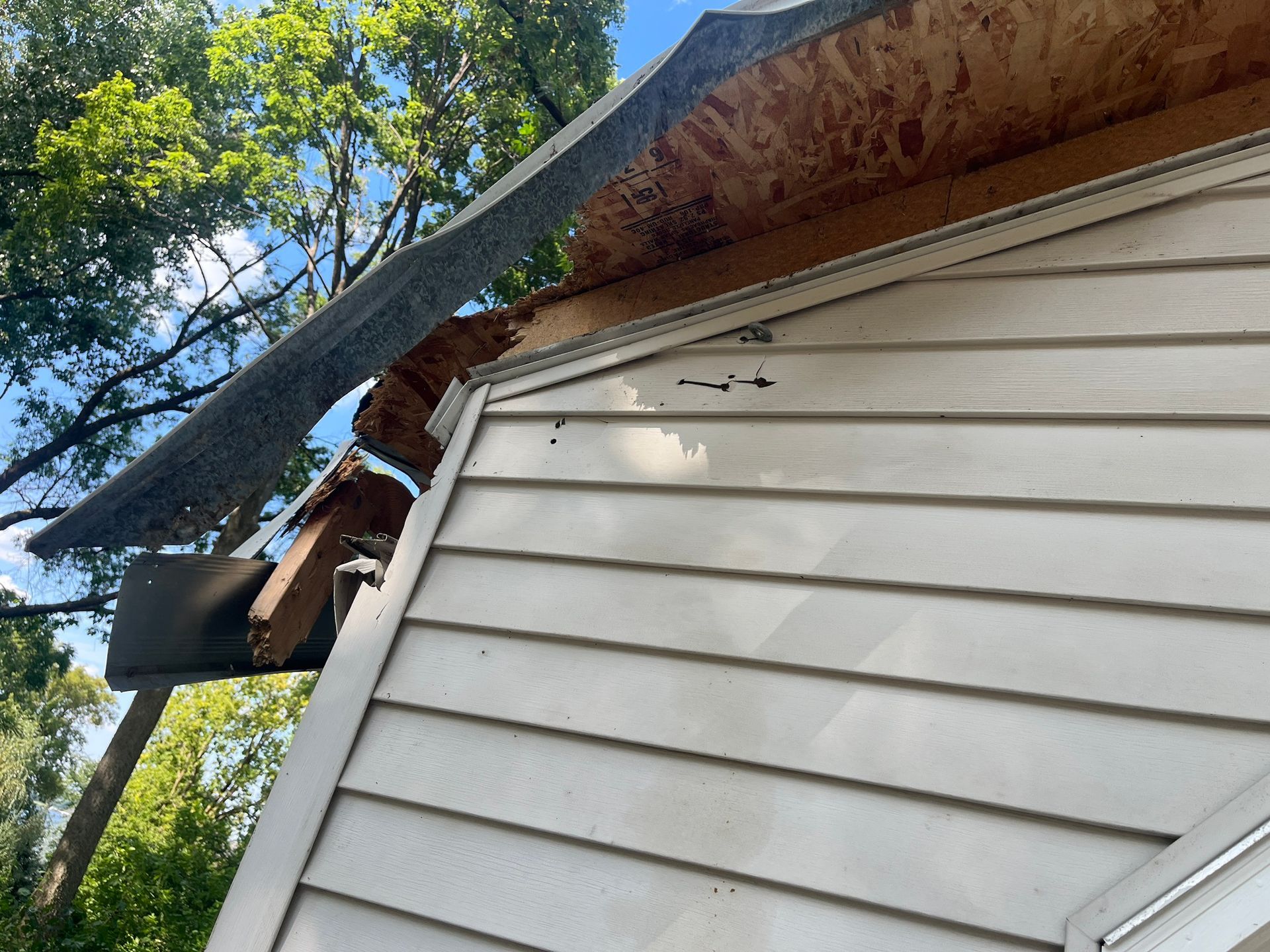 Damaged roof with broken shingles and exposed wood above white siding under trees
