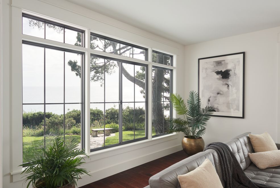 Bright living room with large windows, green view, and a gray sofa beside a potted plant and wall art