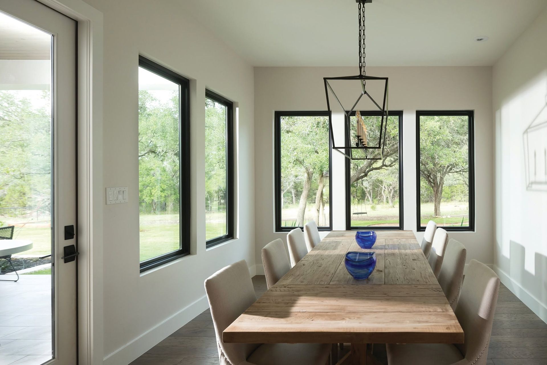 Bright dining room with a wooden table, chairs, black-framed windows, and a glass pendant light