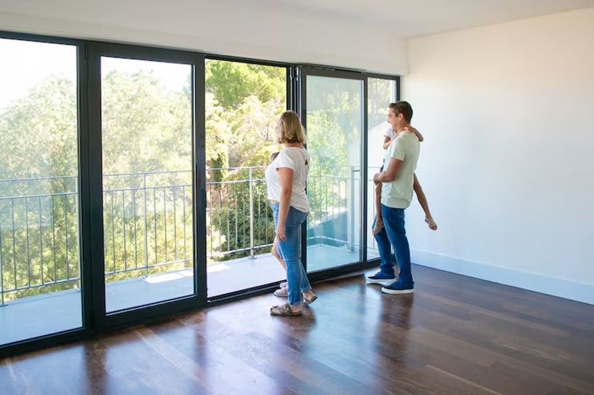 Two people standing in an empty room with large windows and dark wood floors, looking outside.