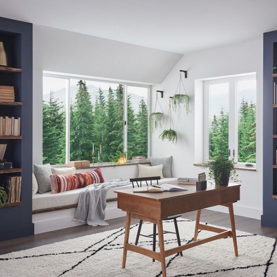 Bright modern living room with large windows, indoor plants, a wooden desk, and a cushioned window seat.